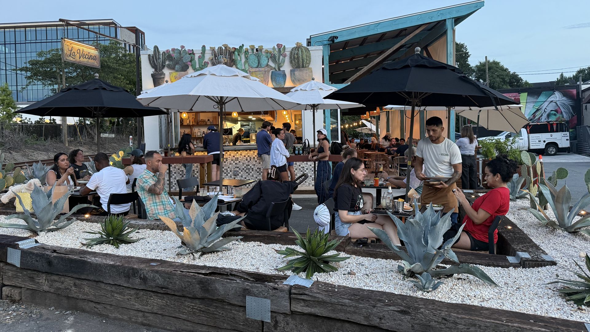 Outdoor dining scene with many people beneath white and black umbrellas, a La Vecina sign on the left, a colorful cactus mural, foreground succulents, and a teal-roofed shelter under a clear blue sky.