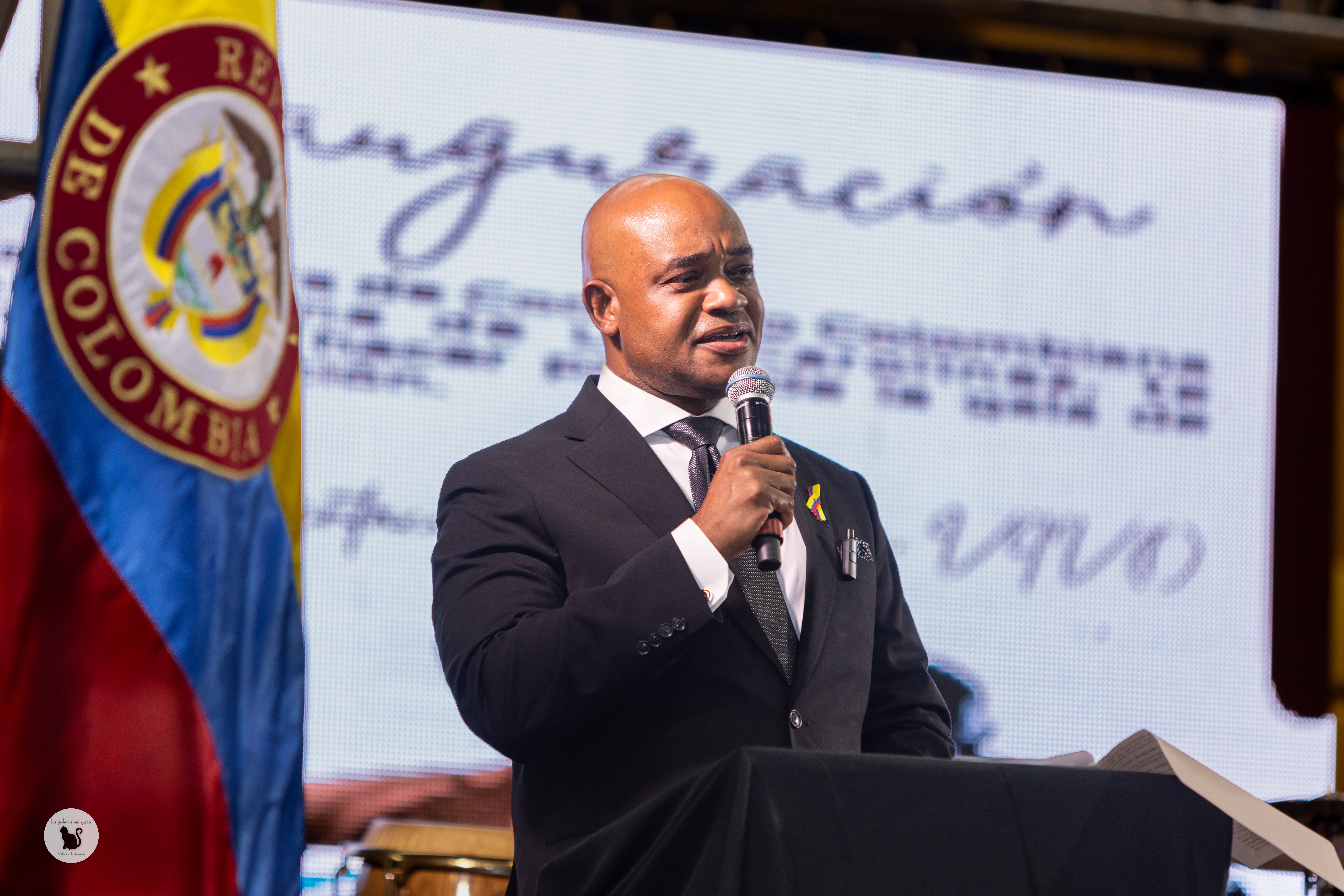 A man in a suit and tie is speaking into a microphone at a podium. He wears a pin with a small Colombian flag on his lapel. Behind him, there is a large display screen featuring the Colombian emblem and some text. The Colombian flag is visible to the left of the image.