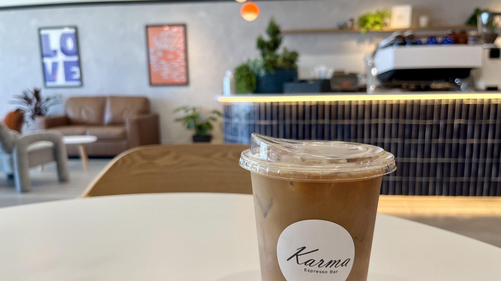 A close-up view of iced coffee in a clear to-go cup with a white sticker of a Karma Espresso Bar logo on it. It sits on a white, rounded table with a blurred cafe in the background showing a blue-tiled bar, plants, and framed art.