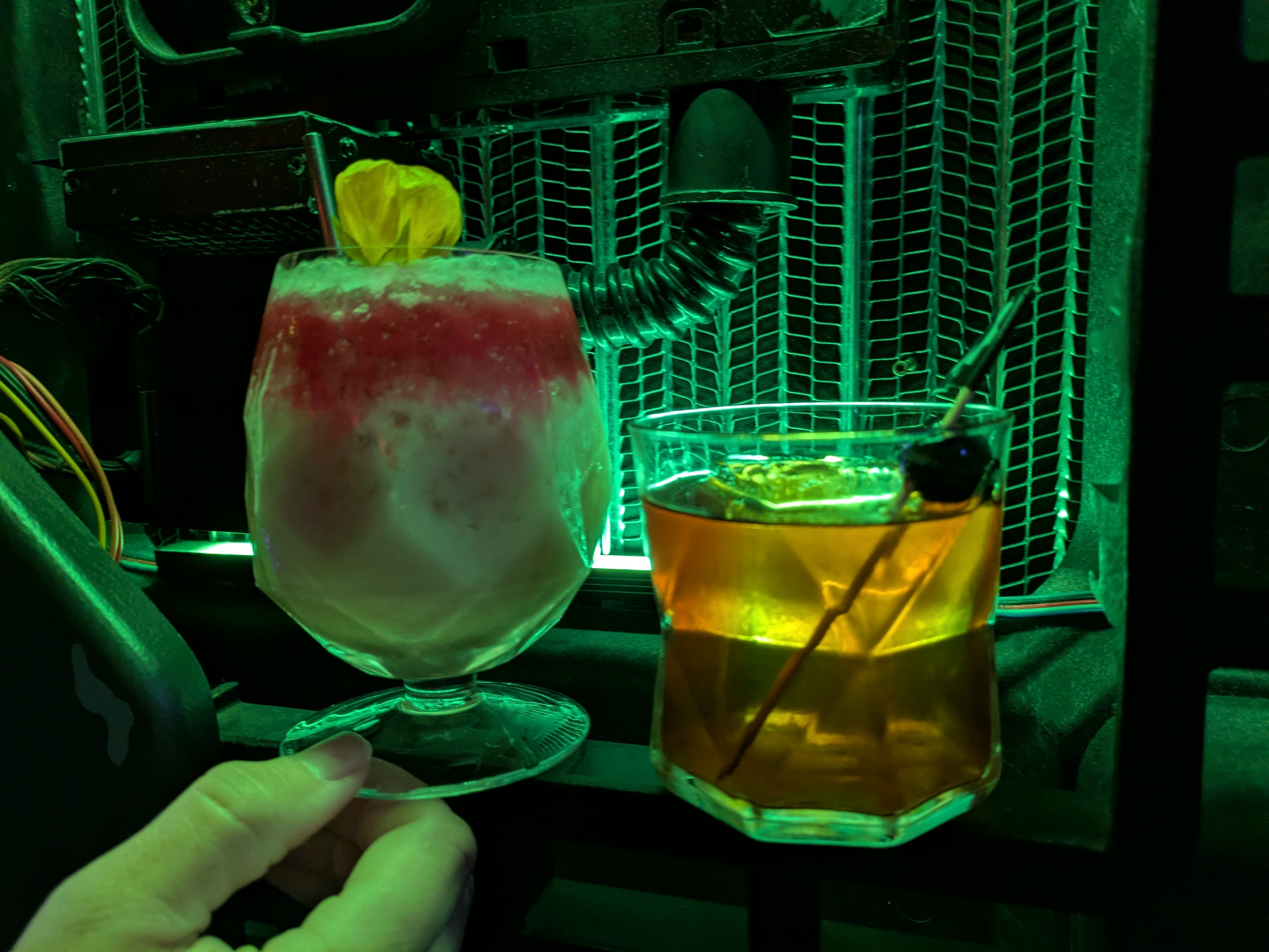 Two cocktails in a dim green-lit setting: a layered pink and white drink garnished with a yellow flower held in a hand, and a brown drink in a short glass with a cherry on a stick.