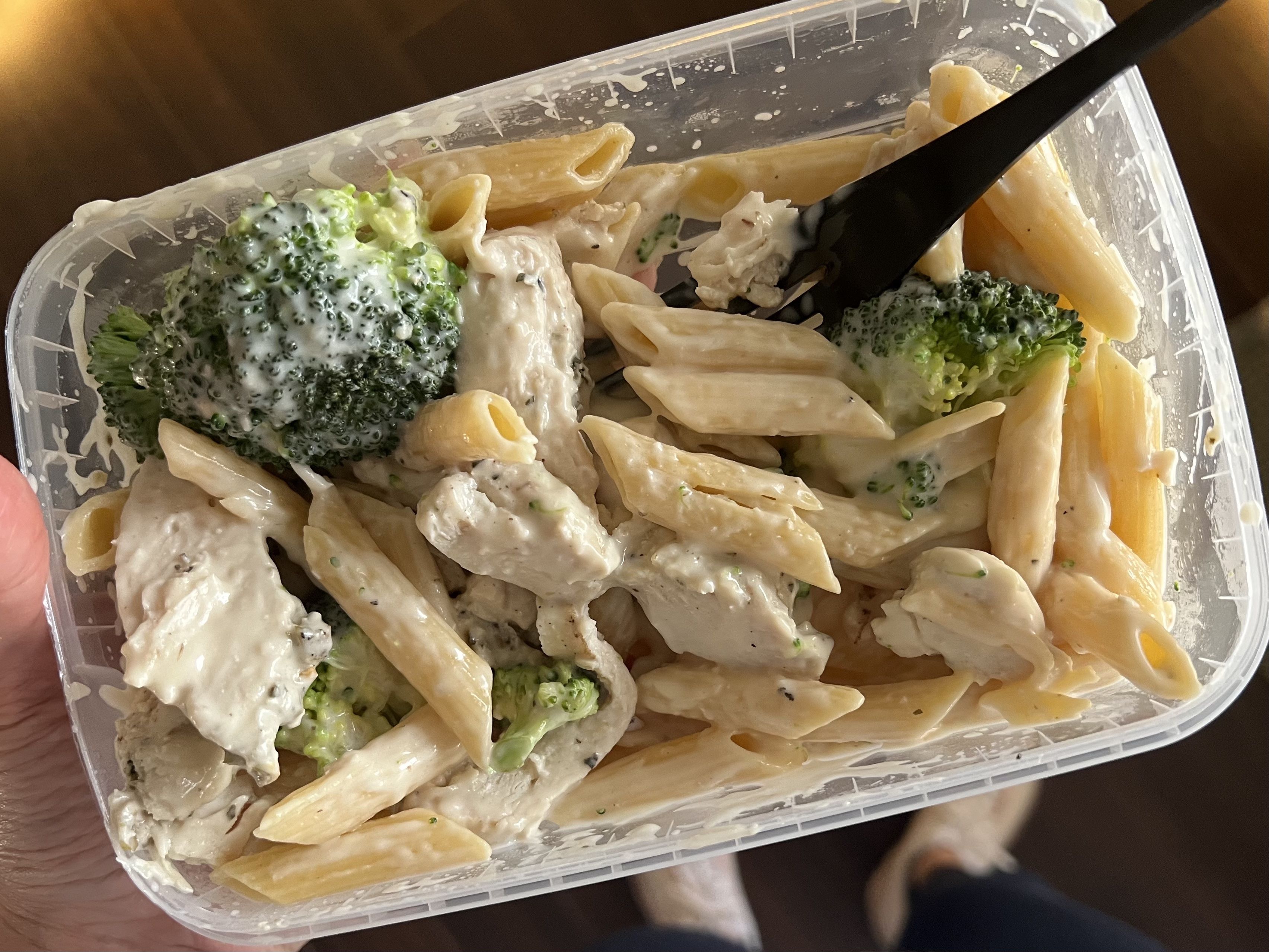 Takeout container with creamy penne pasta, broccoli florets, and chunks of chicken, held by a hand with a black plastic fork inside.