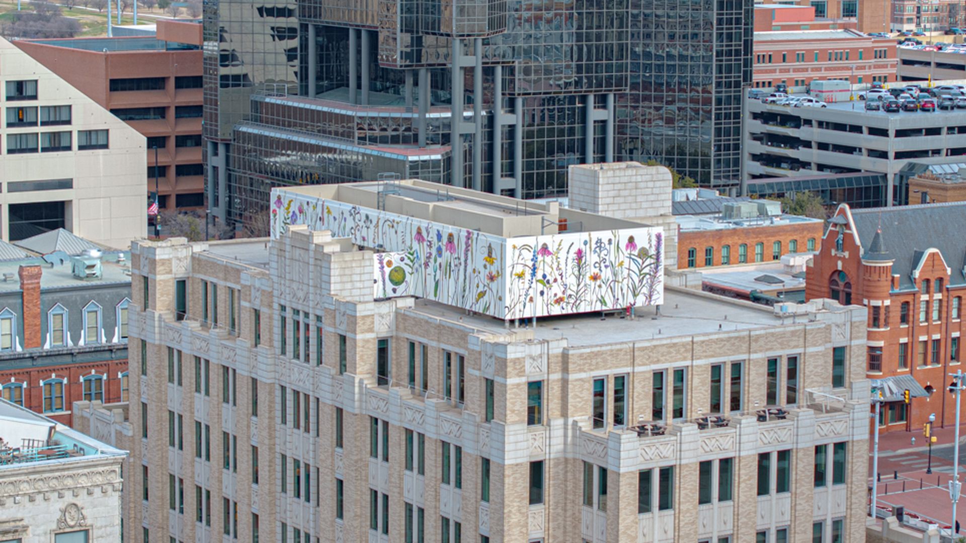 Cityscape with a beige stone building featuring colorful floral murals on rooftop panels, surrounded by modern glass and red brick buildings under a cloudy sky.
