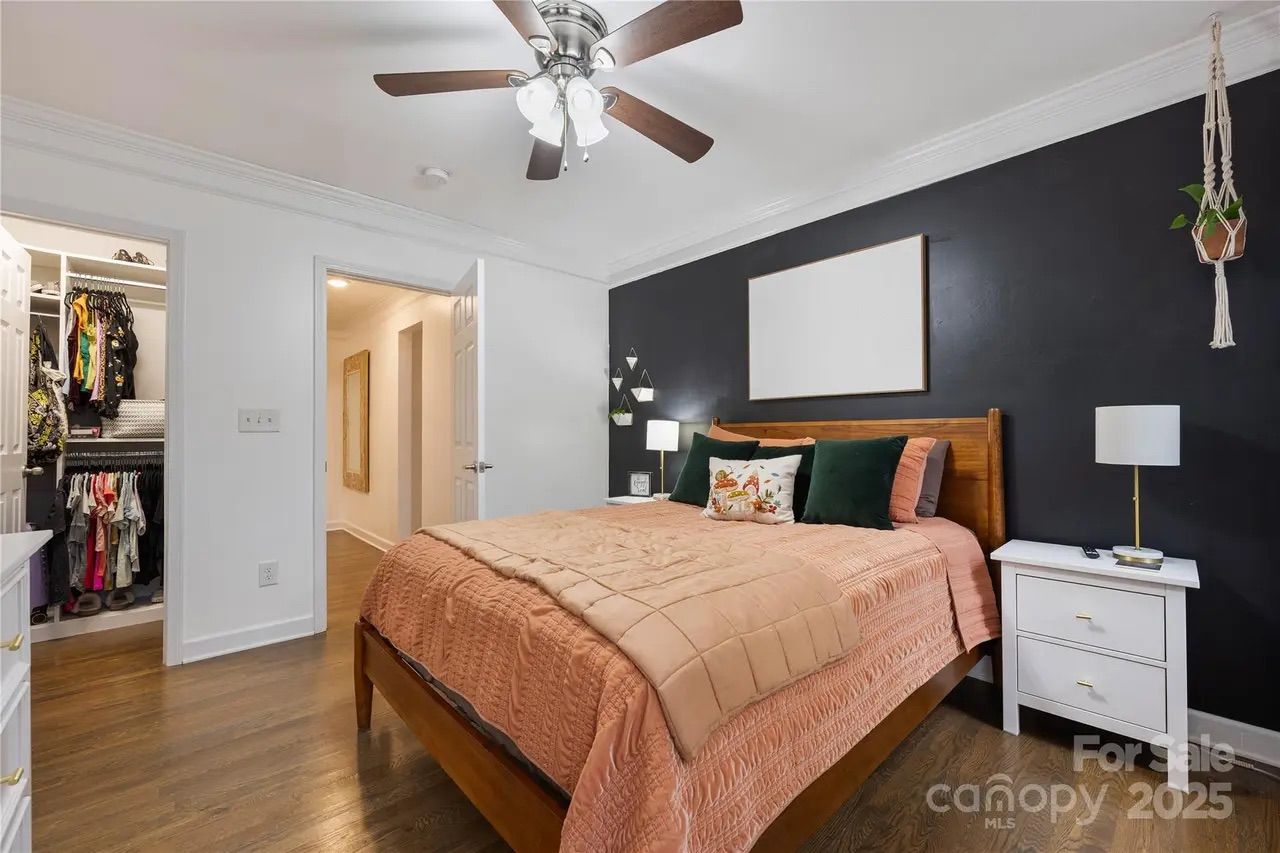 Bedroom with wooden bed frame, peach quilt, dark green pillows, black accent wall with blank art, white bedside tables, hanging plant, ceiling fan, and open doorway to walk-in closet.