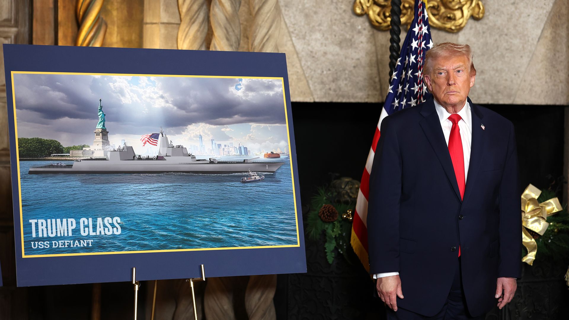 President Trump, in a dark suit and red tie, stands beside a large blue poster showing the Statue of Liberty near a battleship.