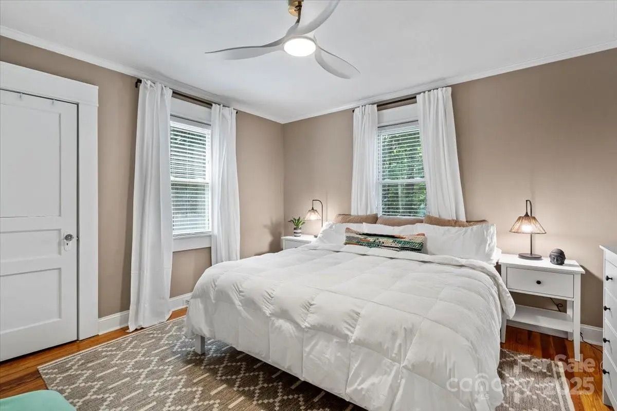 Cozy bedroom with beige walls, white ceiling fan, king-size bed with white comforter, white curtains on two windows, wooden floor, patterned rug, and white nightstands with lamps.