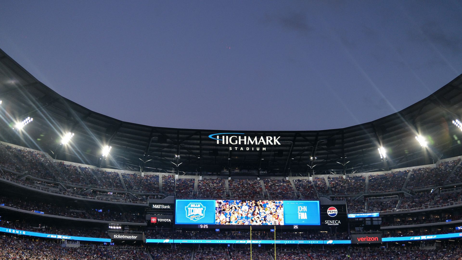 Wide stadium interior at dusk with crowd-filled stands, bright floodlights, a curved roof, and a prominent "HIGHMARK STADIUM" sign; big screen shows fans, field below.