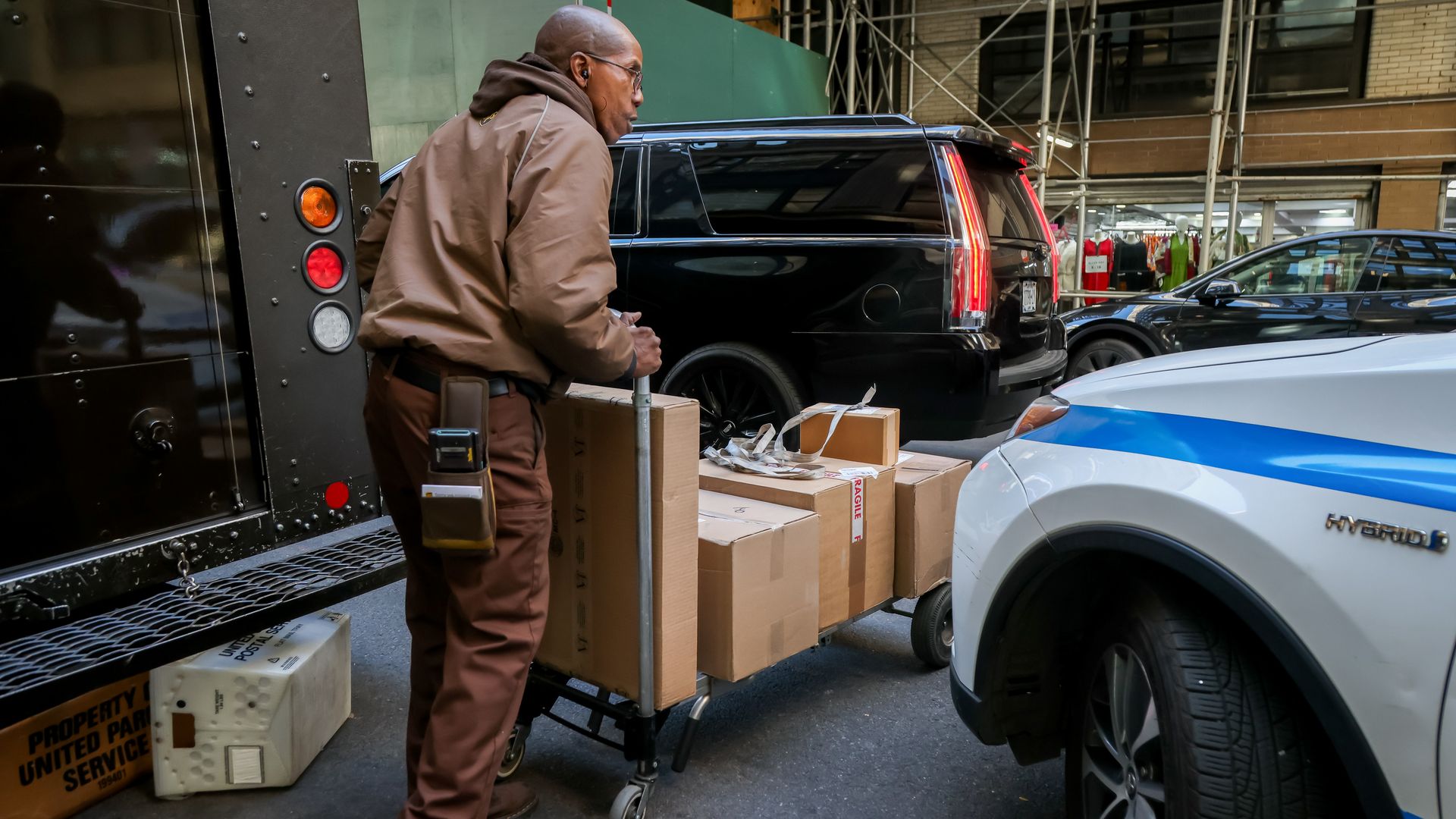Brown-uniformed delivery worker pushes a box-filled cart beside a black SUV and a white police SUV on a city street, with several packages nearby.