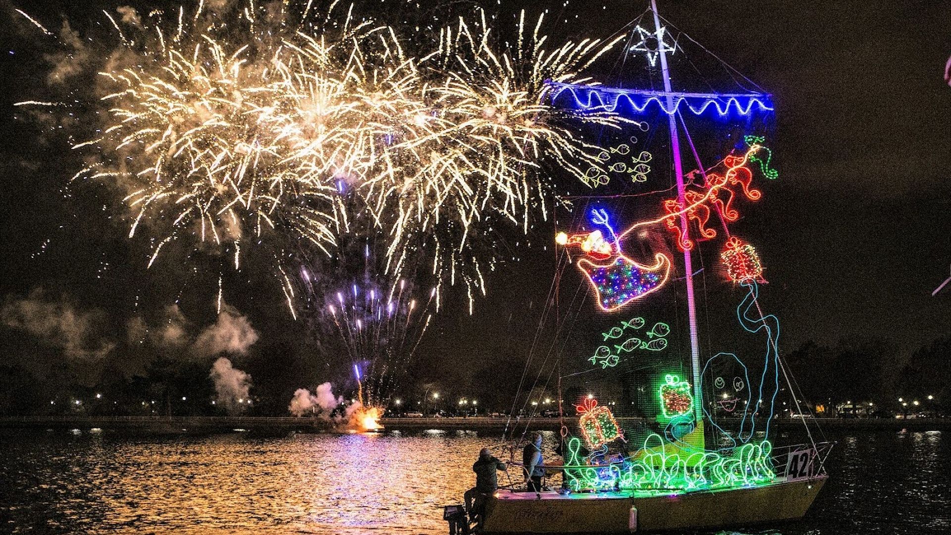 A small boat decorated in holiday lights sailing on water underneath a sky full of fireworks.