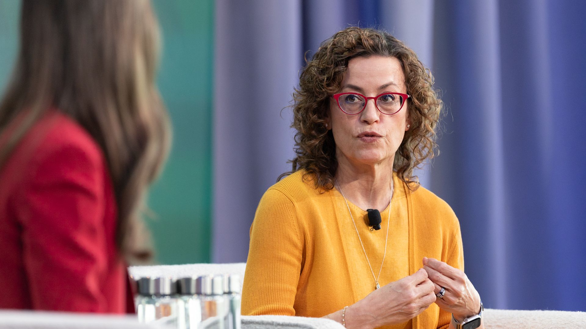 Portrait of a woman with curly brown hair and red glasses, wearing a mustard yellow top, speaking in a TV studio. A lapel mic is clipped to her shirt; blue curtains behind and a blurred person in red on the left.