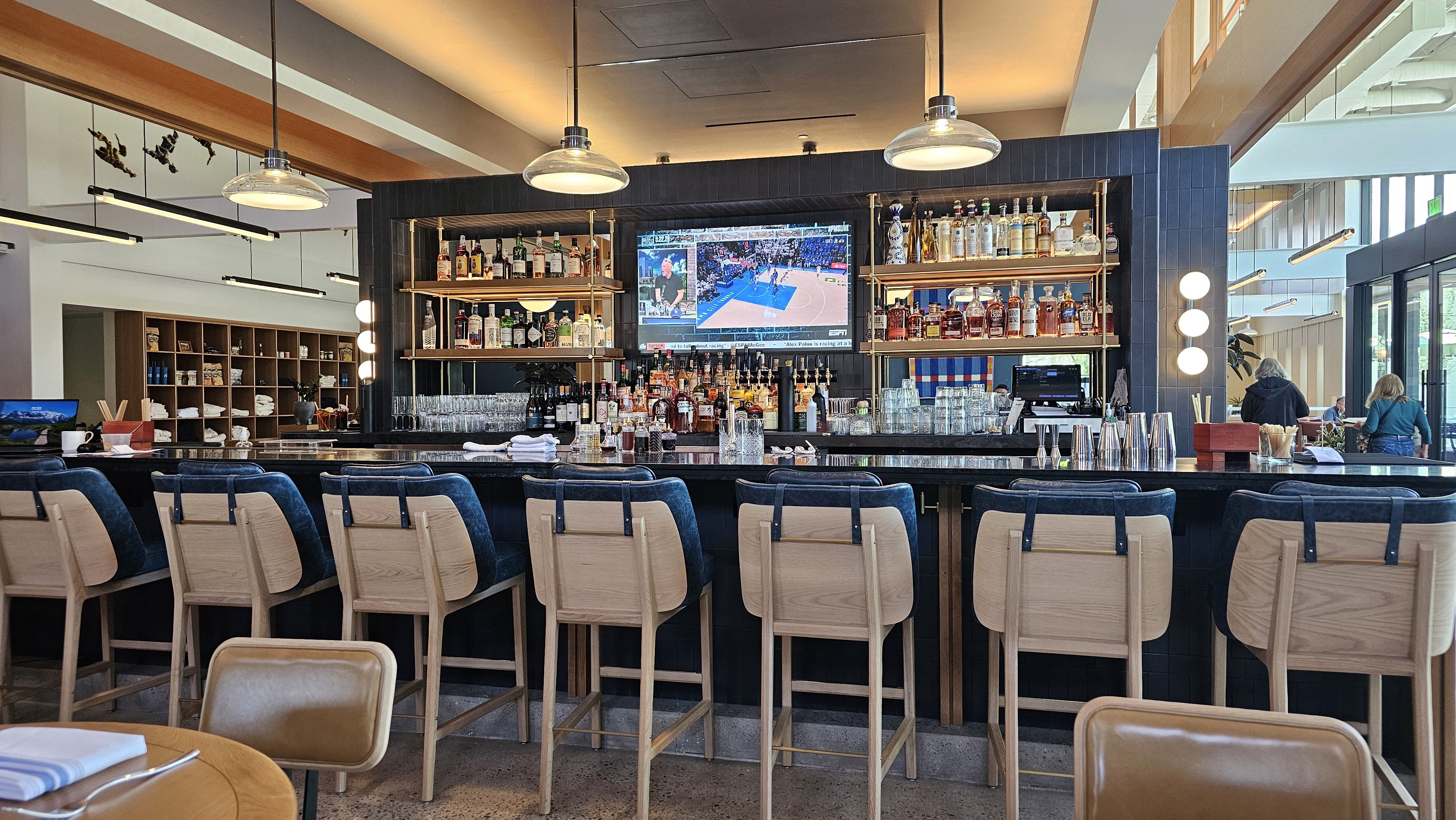 Bright modern bar with navy tile wall, copper shelves of bottles, a long dark counter with wooden stools, a mounted TV showing a basketball game, warm glass dome lights.