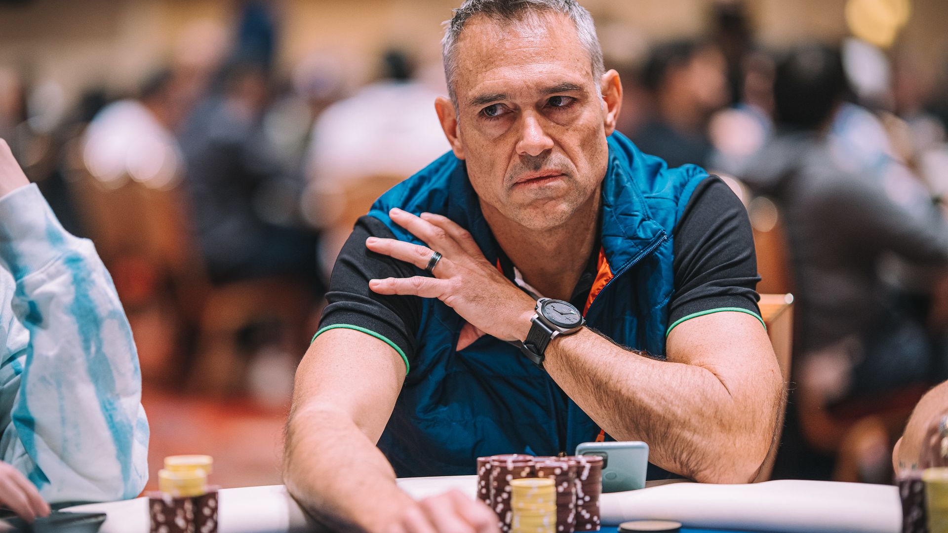 Graying man in a blue vest sits at a poker table, glancing to the side with a tense expression. Chip stacks in front; blurred players in a busy room behind him.