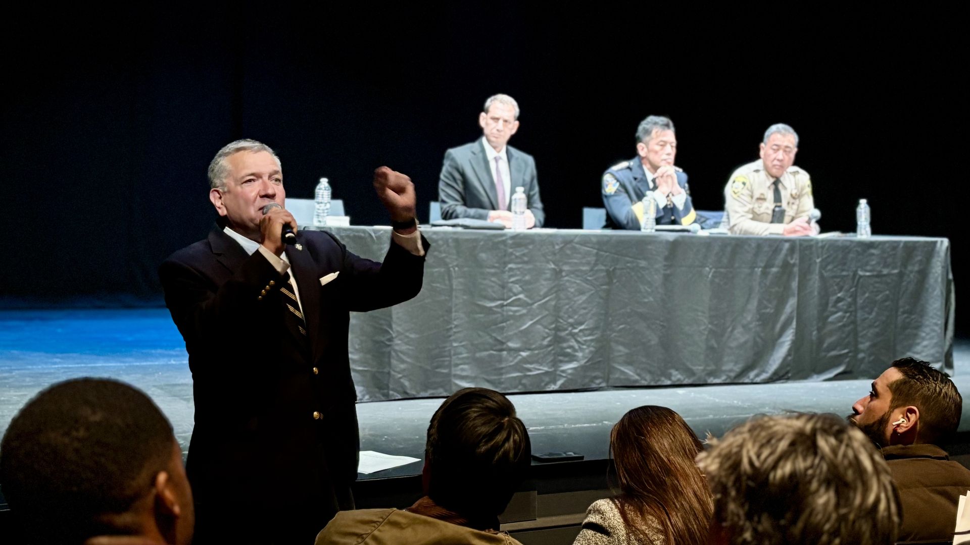 Man in a dark suit speaking into a microphone on stage, gesturing with his hand, while three men in suits and uniforms sit at a table with water bottles behind him, audience in foreground.