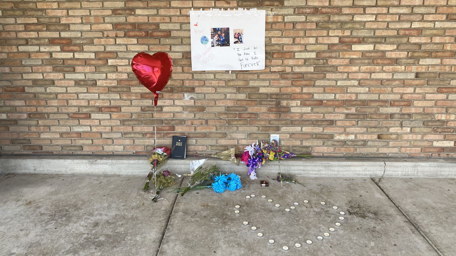 A memorial for Ta'Kiya Young on the side of a Kroger building, with a poster, heart-shaped balloon, flowers and tea light candles shaped in a heart