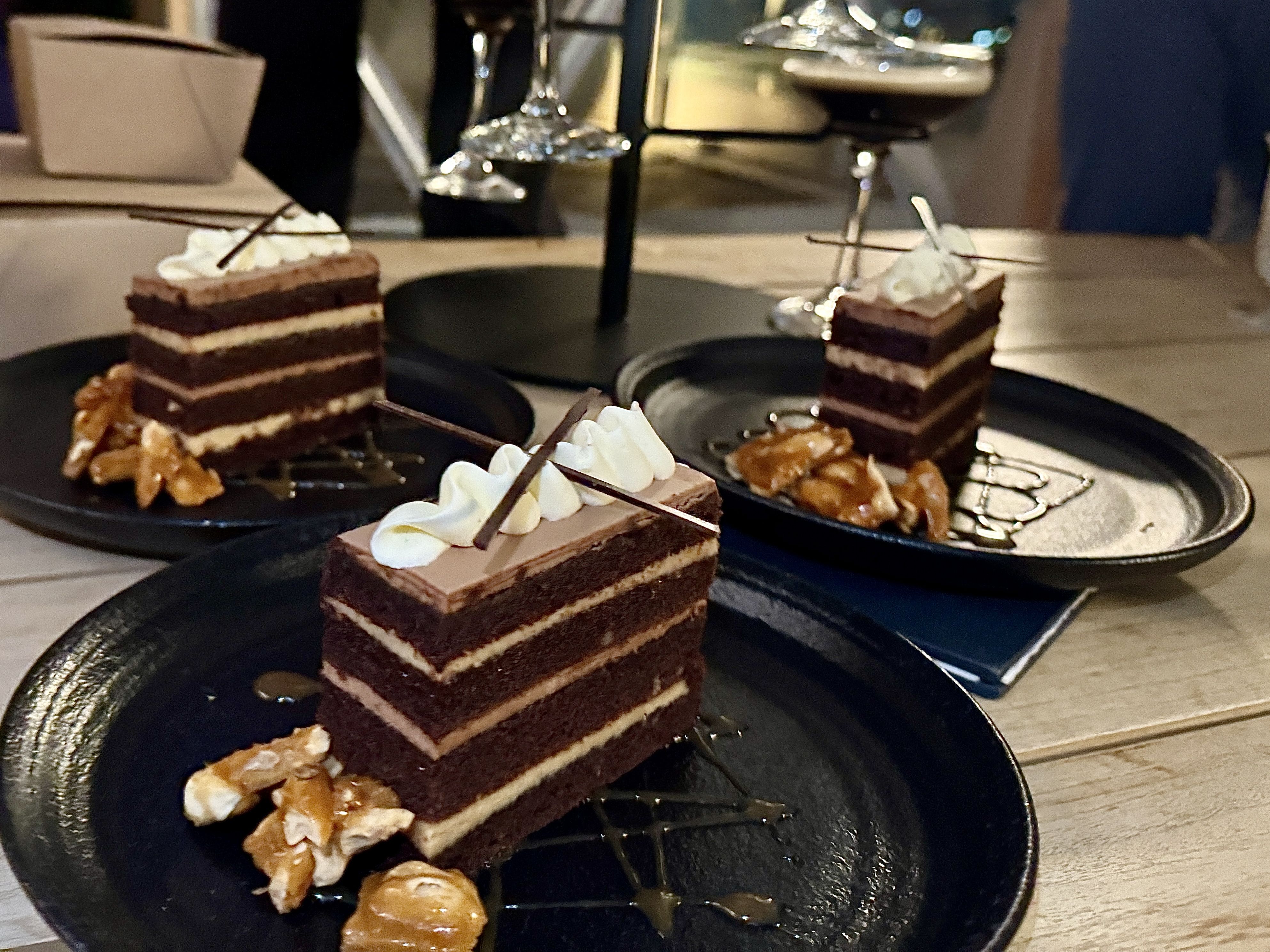 Three slices of layered chocolate cake with cream topping and chocolate garnish on black plates, each accompanied by caramelized nuts, set on a wooden table with glassware in the background.