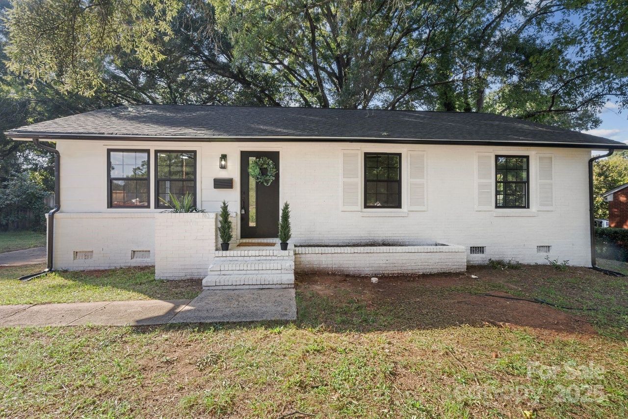 Single-story white brick house with black trim, a black front door with a green wreath, two potted plants by the steps, and large trees in the background.