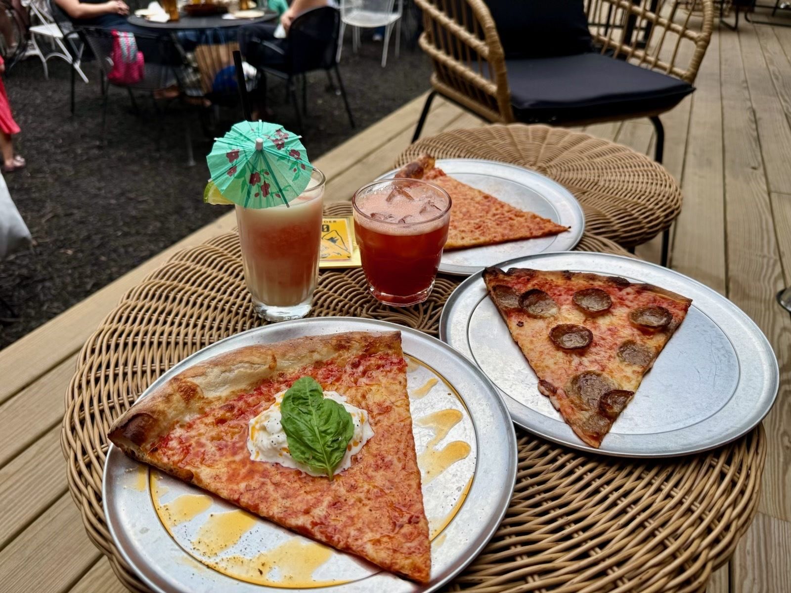 Outdoor wicker table with two pizza slices on metal plates, one topped with a basil leaf; the other with pepperoni; two drinks with small umbrellas; patio chairs in a casual outdoor setting.
