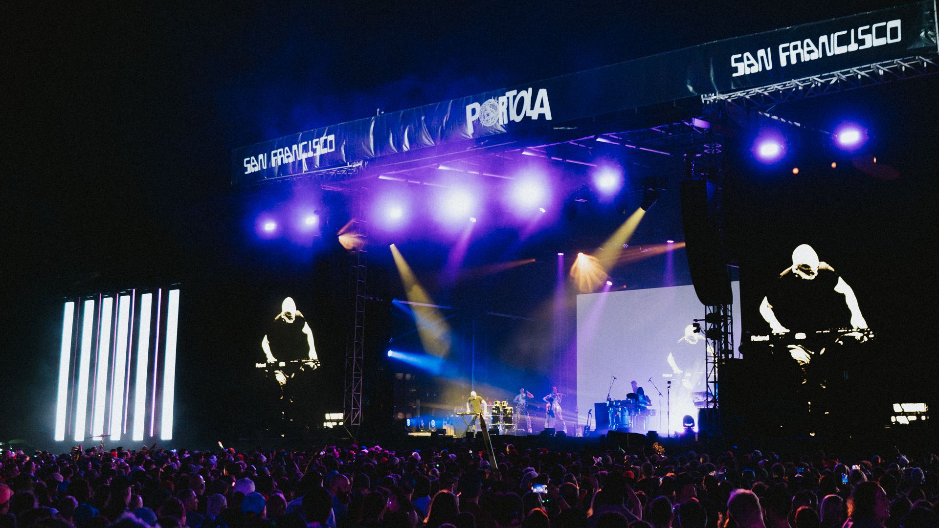 Nighttime outdoor concert with a large crowd in front of a bright stage. Purple and blue lights beam from the rigging, screens show performers, and banners read "SAN FRANCISCO" and "PORTOLA" above.
