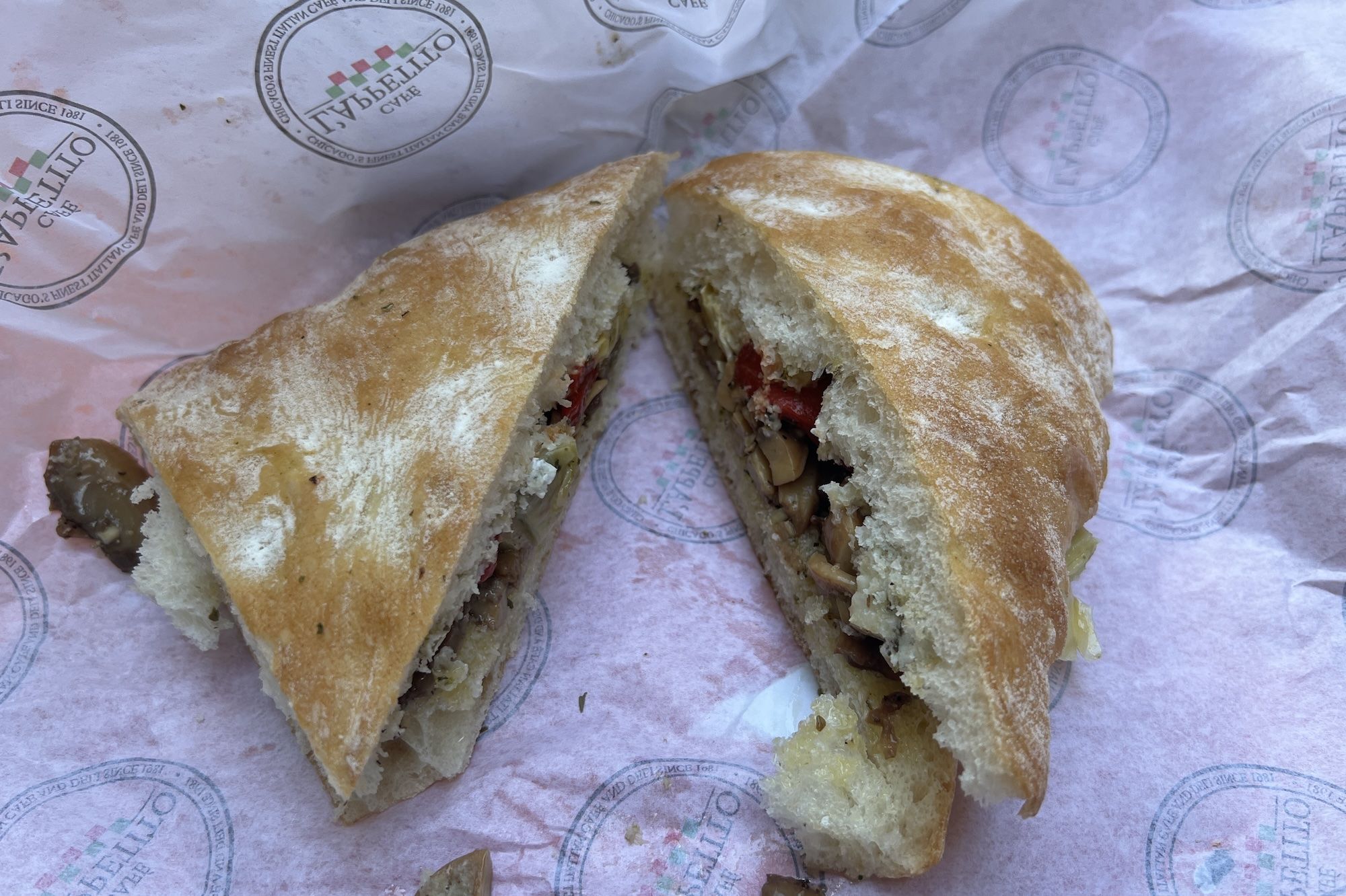 Split crusty white bun filled with sautéed mushrooms, pieces of red pepper and melted cheese; ends tucked inside a pink parchment with circular stamped logos.