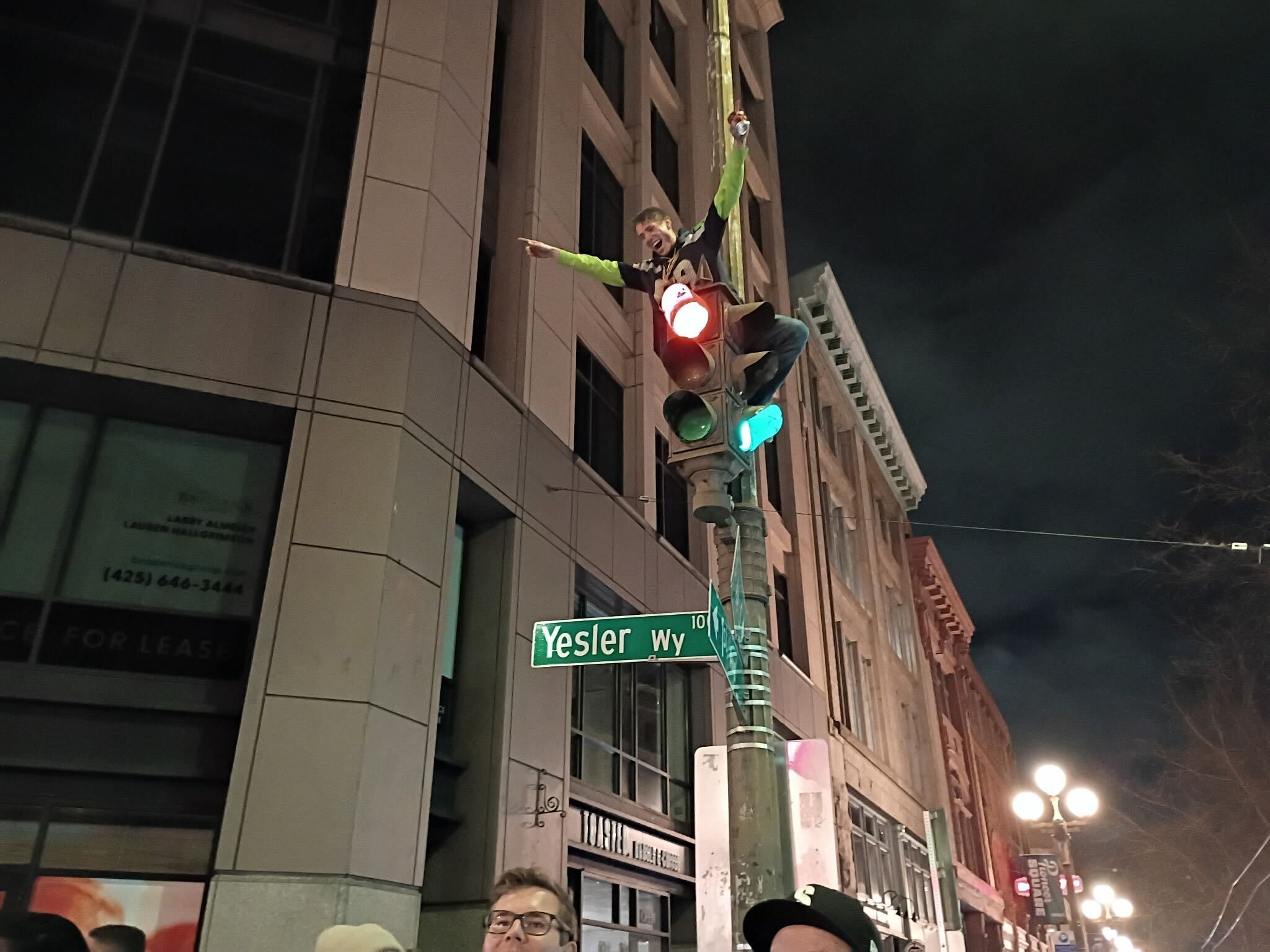 Person in a black and yellow shirt climbing a traffic light pole at night on Yesler Way, smiling and pointing, with buildings and street lamps in the background.