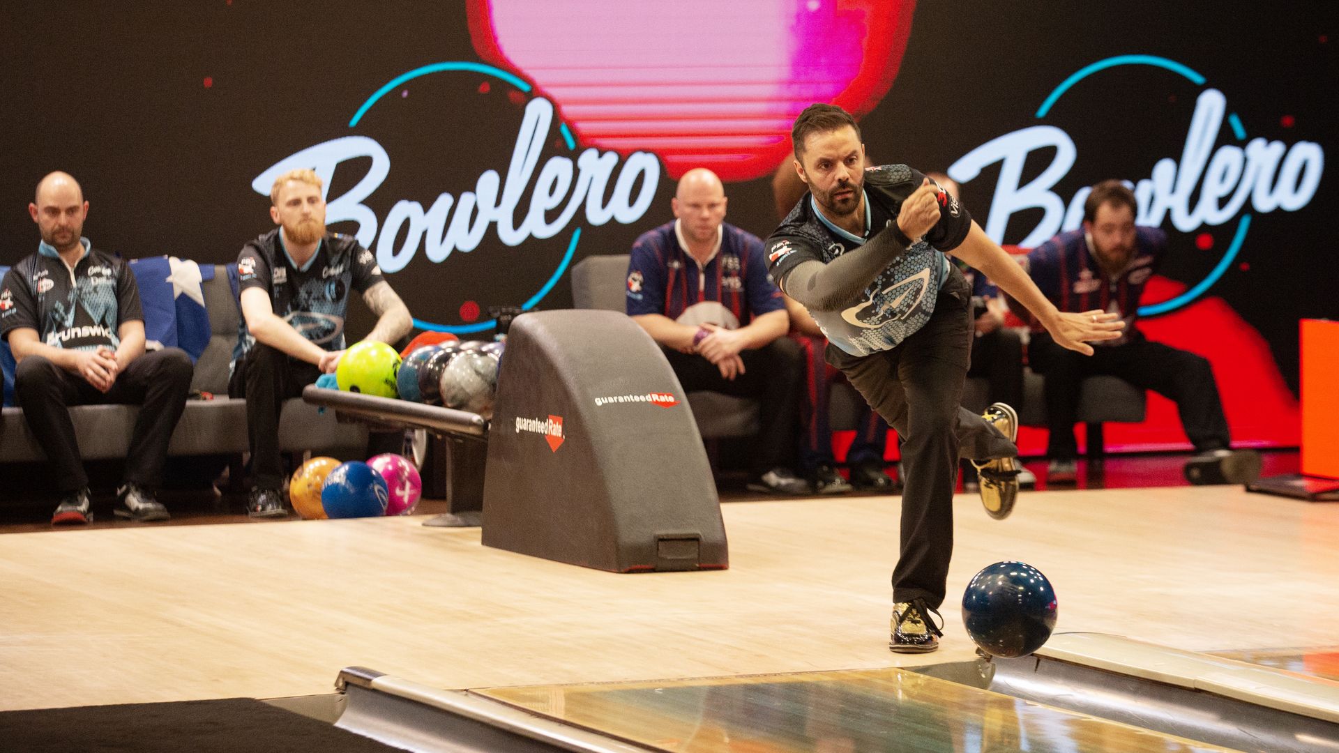 A PBA bowler releases the ball down the lane as competitors look on in the background
