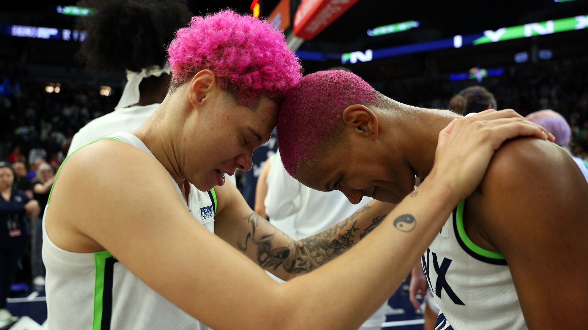 Two basketball players with bright pink hair hug foreheads on the court, wearing white uniforms with green trim. One has visible tattoos; crowd and hoop loom in the blurred background.