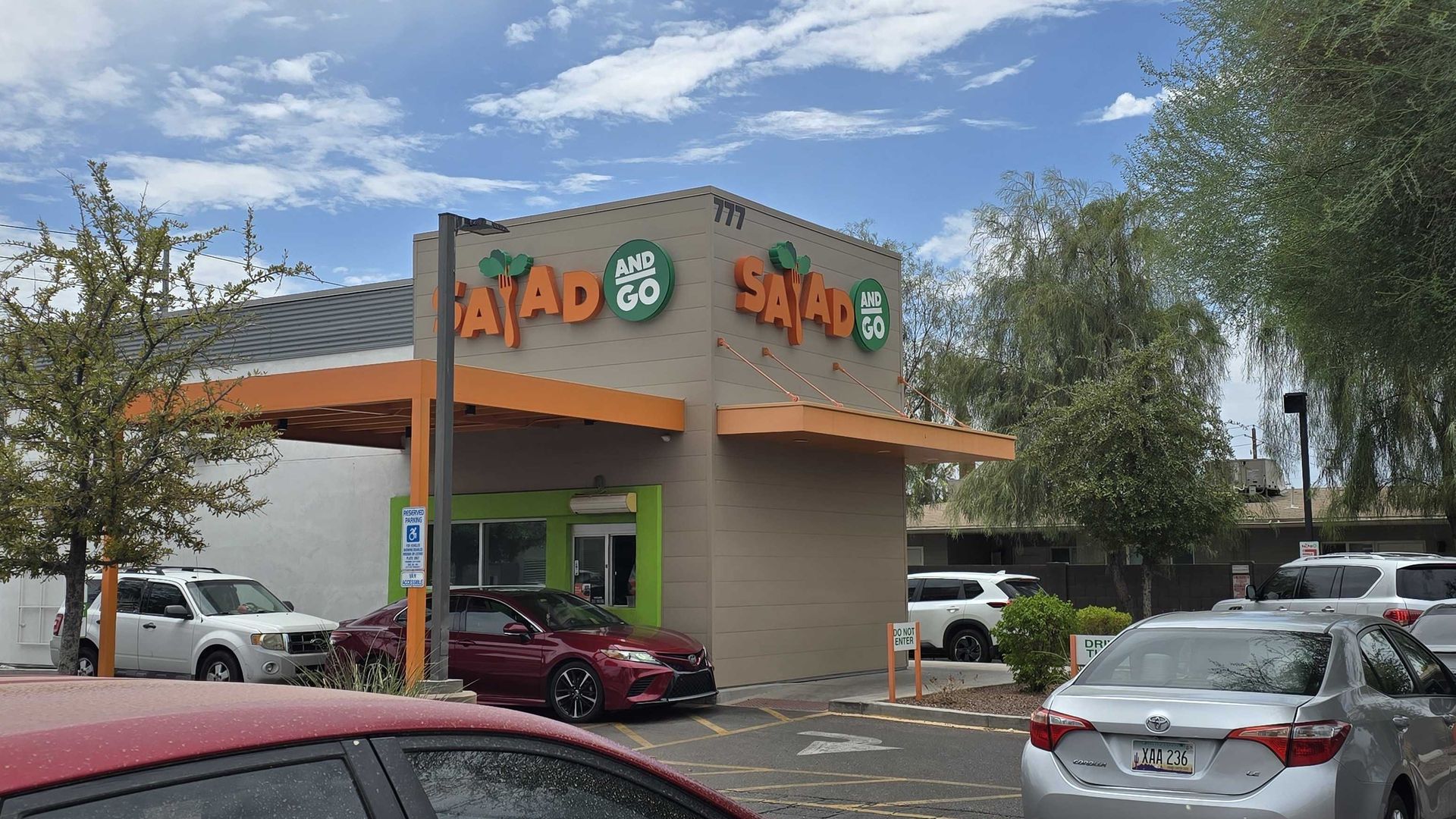 A beige fast-casual restaurant with orange awnings and bold orange lettering "SALAD AND GO"; several cars snake through the drive-thru in a lot with trees and a blue sky.