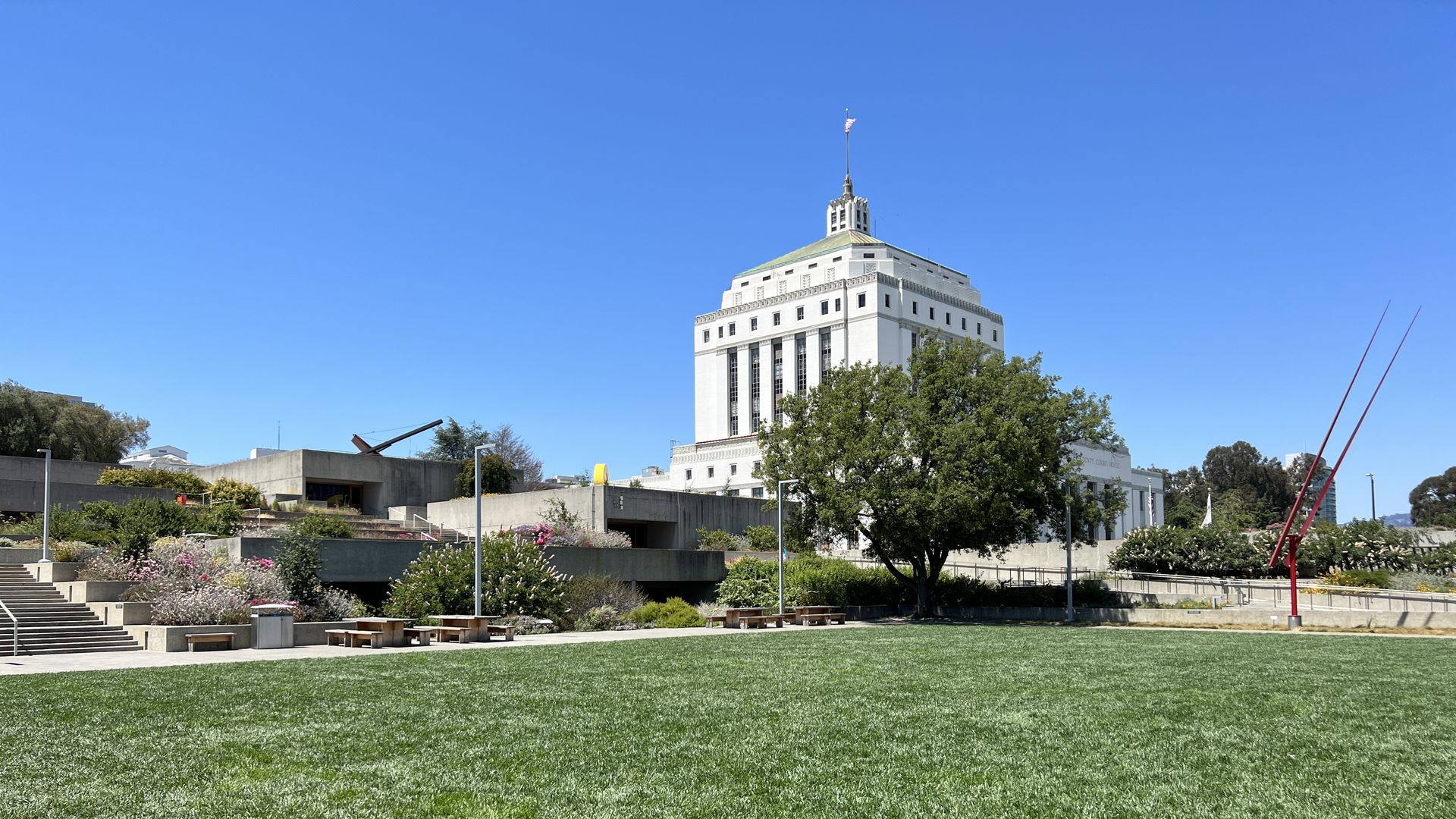 Open green lawn with a tall white building and a large tree in the background under a clear blue sky. Concrete stairs and benches with colorful flowers surround the area.