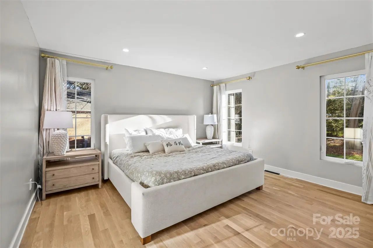 Bright bedroom with light gray walls, wooden floor, large white upholstered bed with pillows including one that says "Mr. & Mrs.", two wooden nightstands with white lamps, and three windows with white curtains.