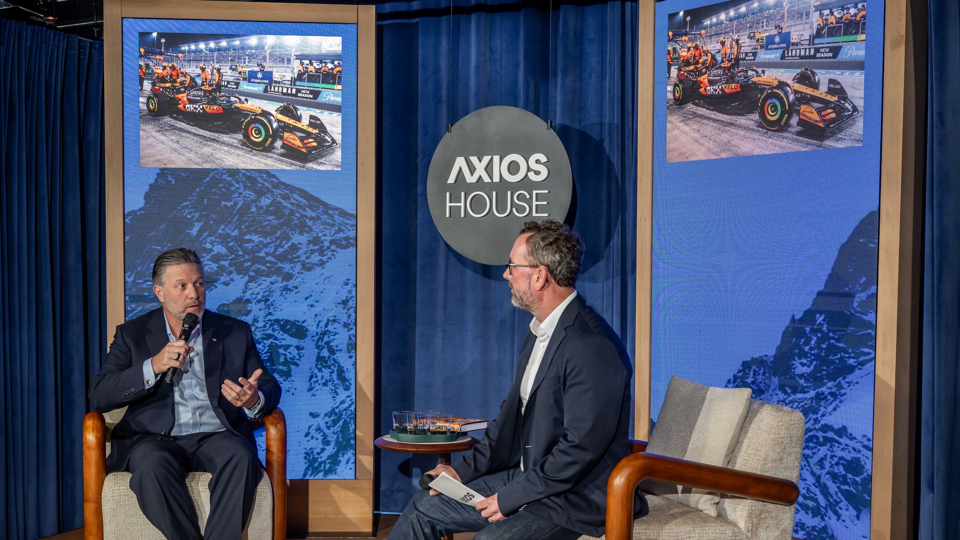 Two men in suits seated on beige armchairs having a discussion on stage with blue curtains and screens displaying race car images, and a sign reading "AXIOS HOUSE" in the center.