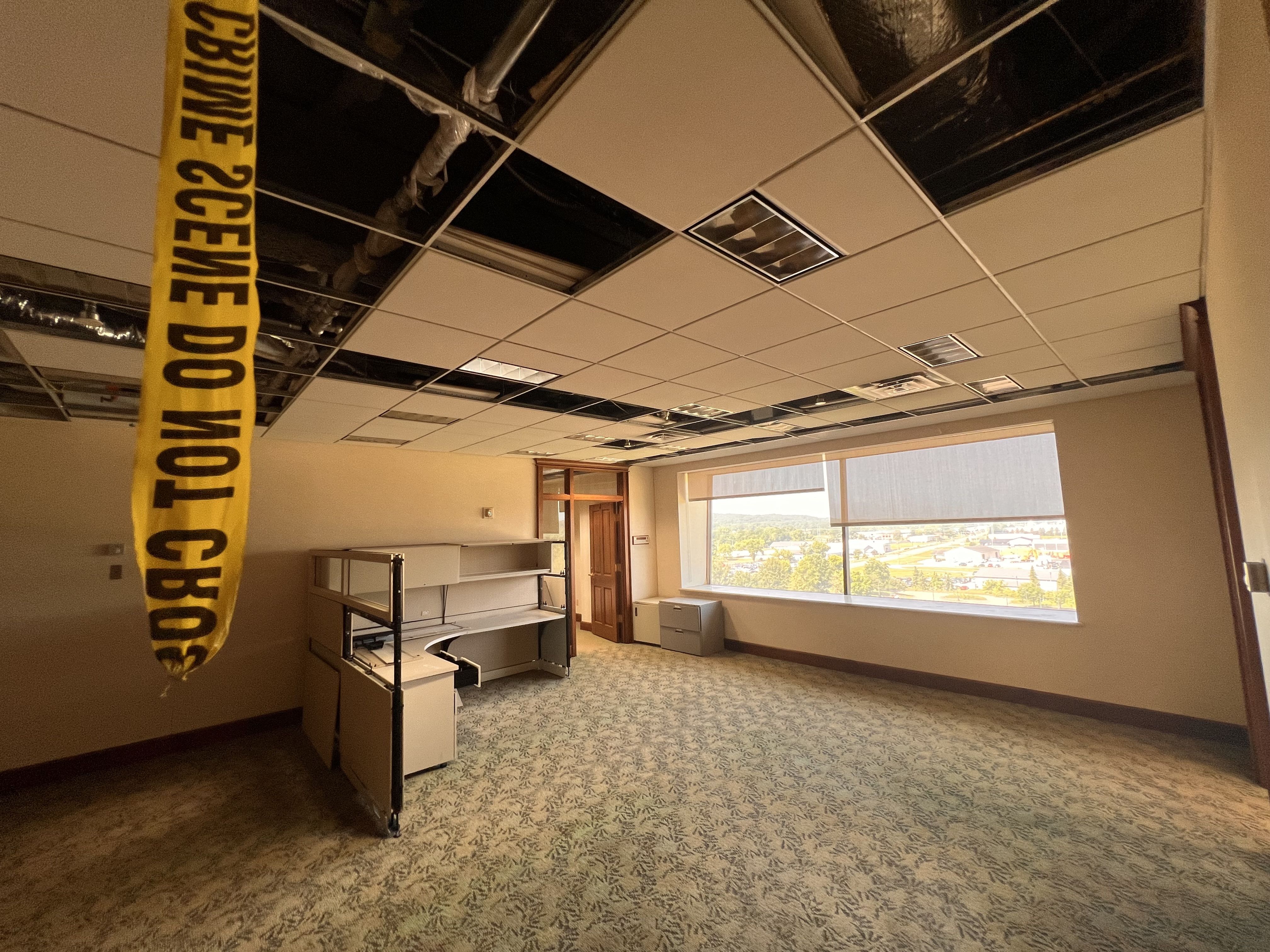 Office interior with a damaged ceiling; several tiles removed to reveal pipes. A yellow caution tape hangs on the left. Empty desks, shelves, and filing cabinets near a large window.