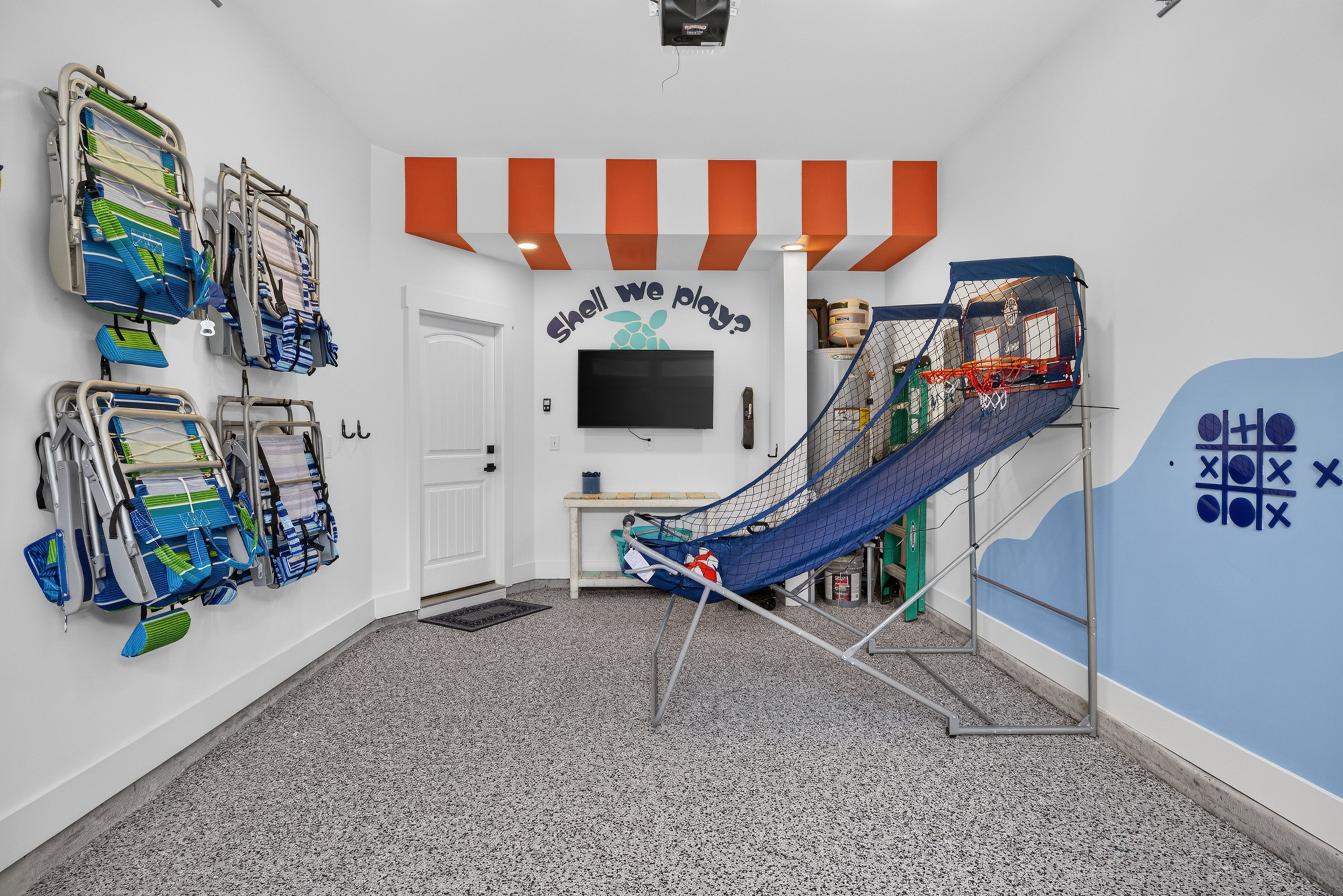 Playroom storage with orange-and-white striped canopy, stacked beach chairs on left, a wall sign reading "Shell we play?" above a TV, and a blue indoor basketball hoop on the right.