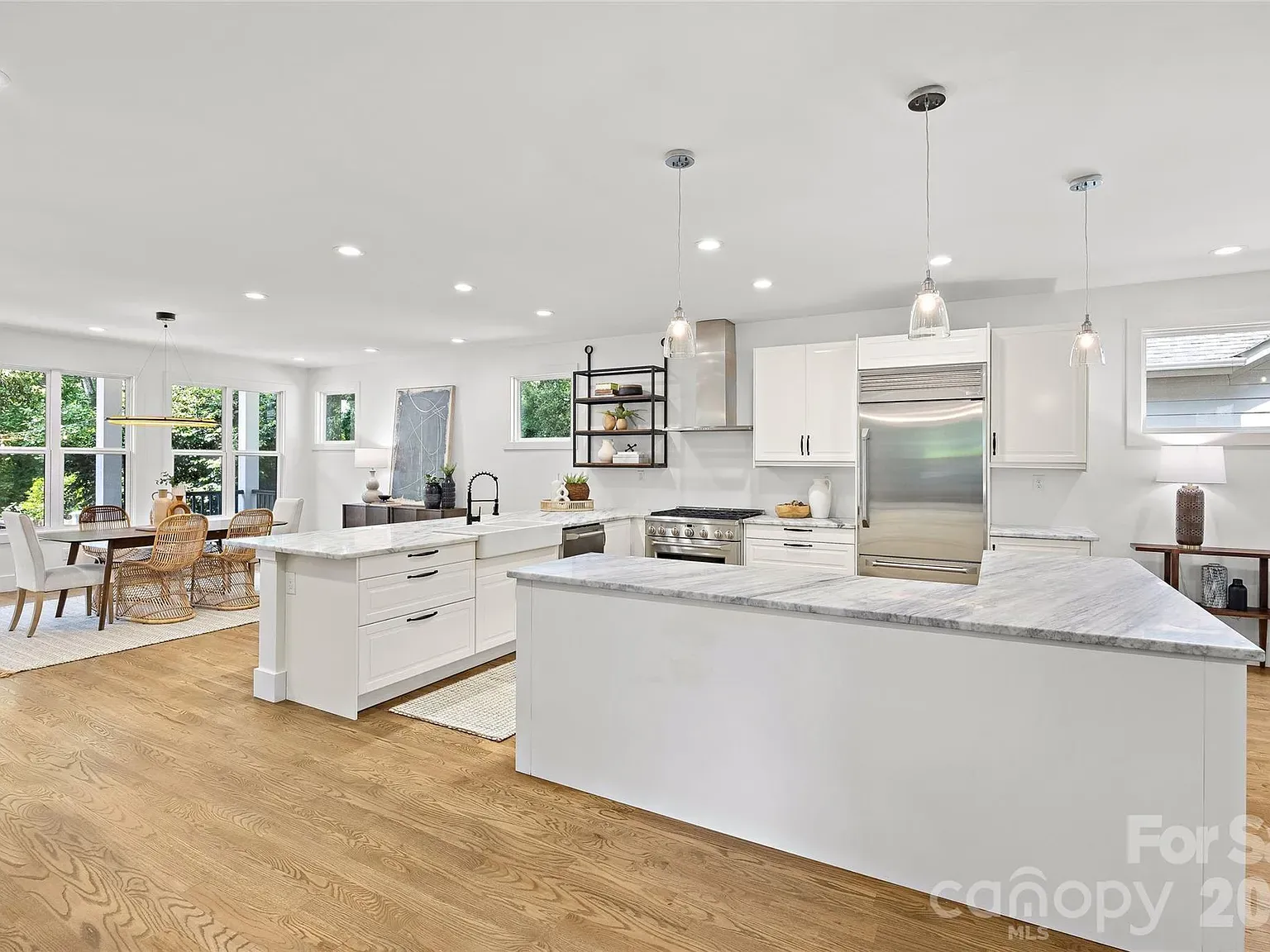 Bright open kitchen with white cabinets and marble countertops, a large island, and stainless steel fridge and range. Dining area with wicker chairs by large windows overlooking greenery.