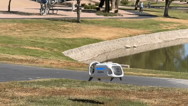 A white, boxy module with a rotor on the back lowers to the ground on a cable, drops a small package and rises into the air. 