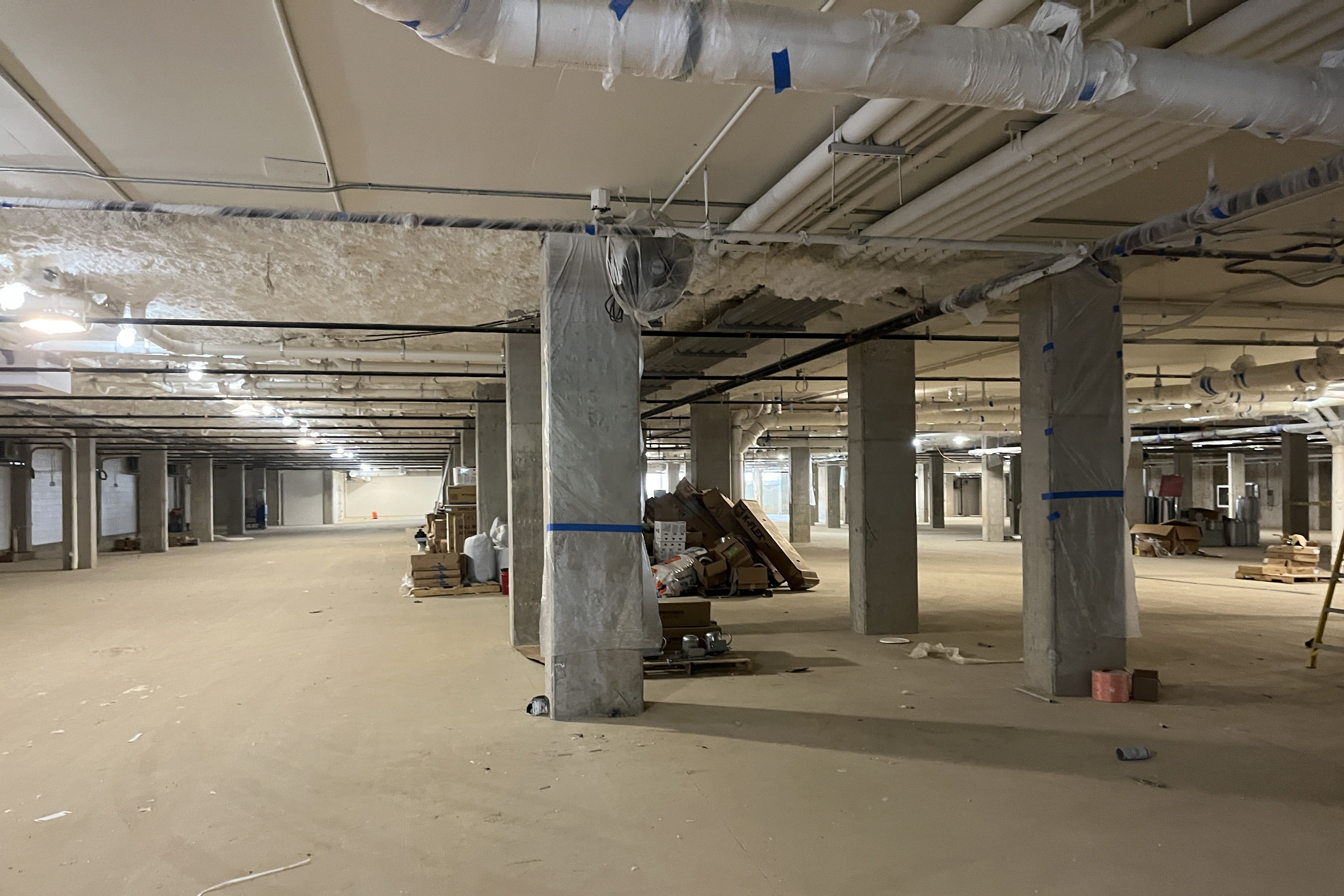 Inside a large unfinished basement under construction: exposed pipes and ducts on the ceiling, wrapped concrete columns, and scattered pallets, boxes, and debris on bare concrete floors.