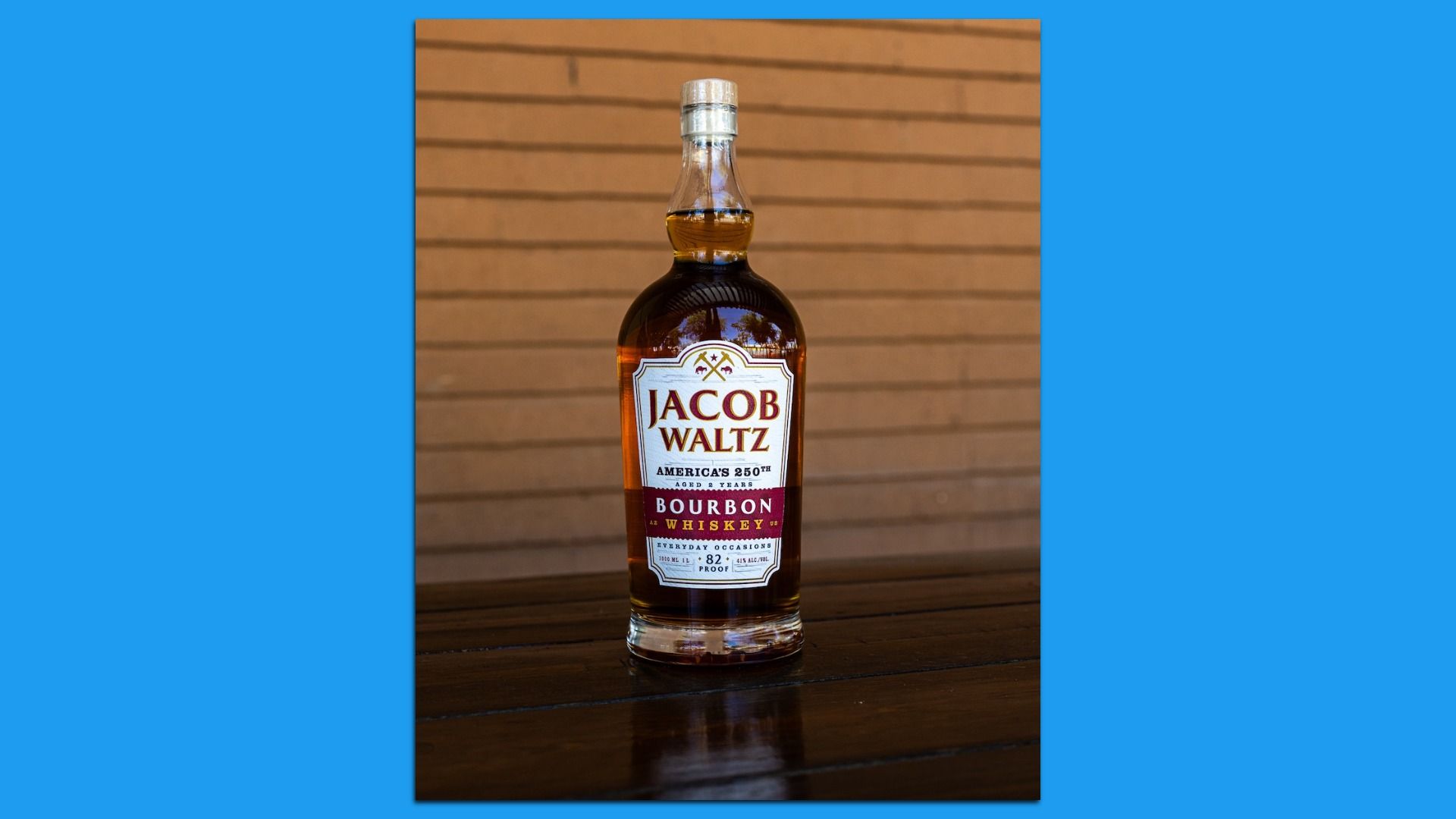 A bottle of Jacob Waltz Bourbon Whiskey sits on a dark wooden table, amber liquid visible. The label reads "JACOB WALTZ" against a brown slat wall.