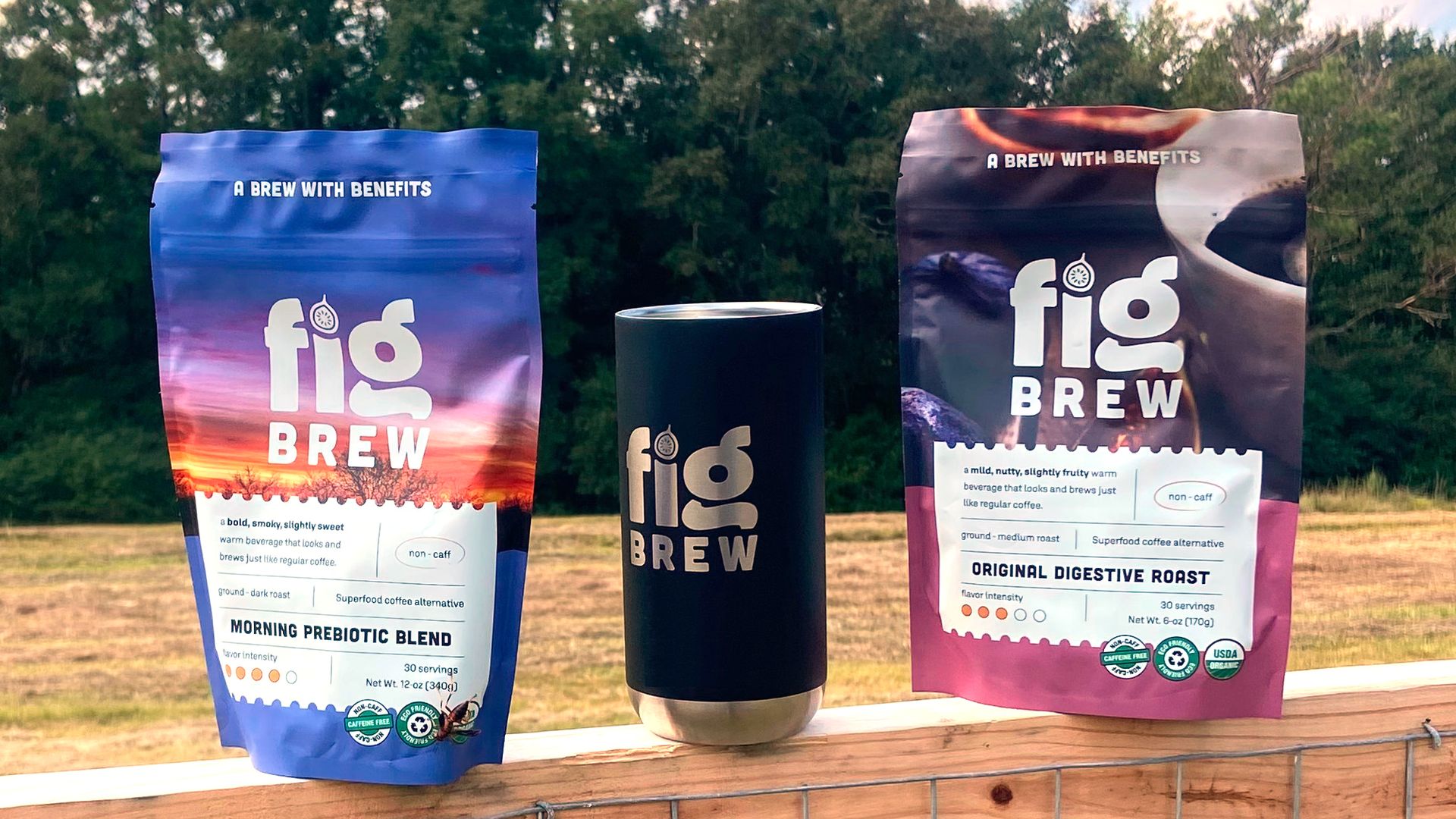 Two colorful Fig Brew packages and a black Fig Brew cup on a wooden fence with green trees and blue sky with clouds in the background.