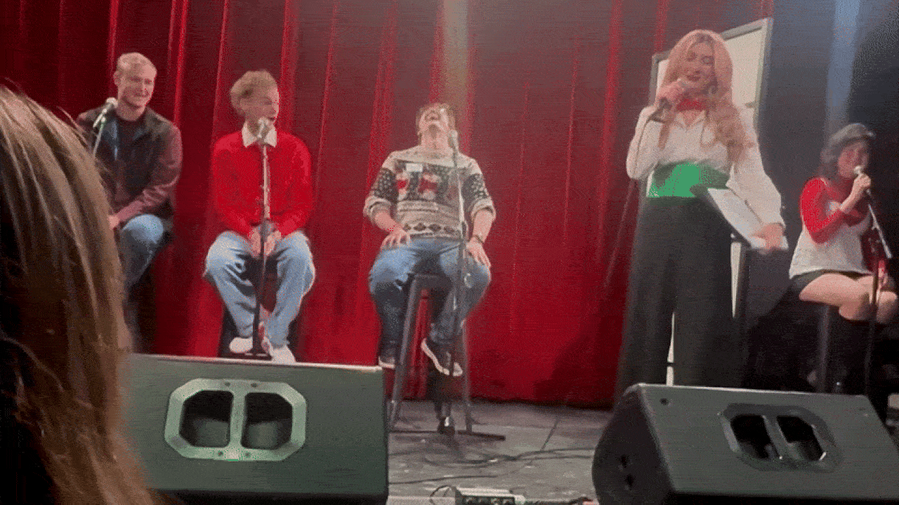 Five people on stage with red curtains backdrop; three sitting on stools with microphones, one in a festive sweater laughing, another in white shirt with green sash singing, and a woman singing seated in red and white.