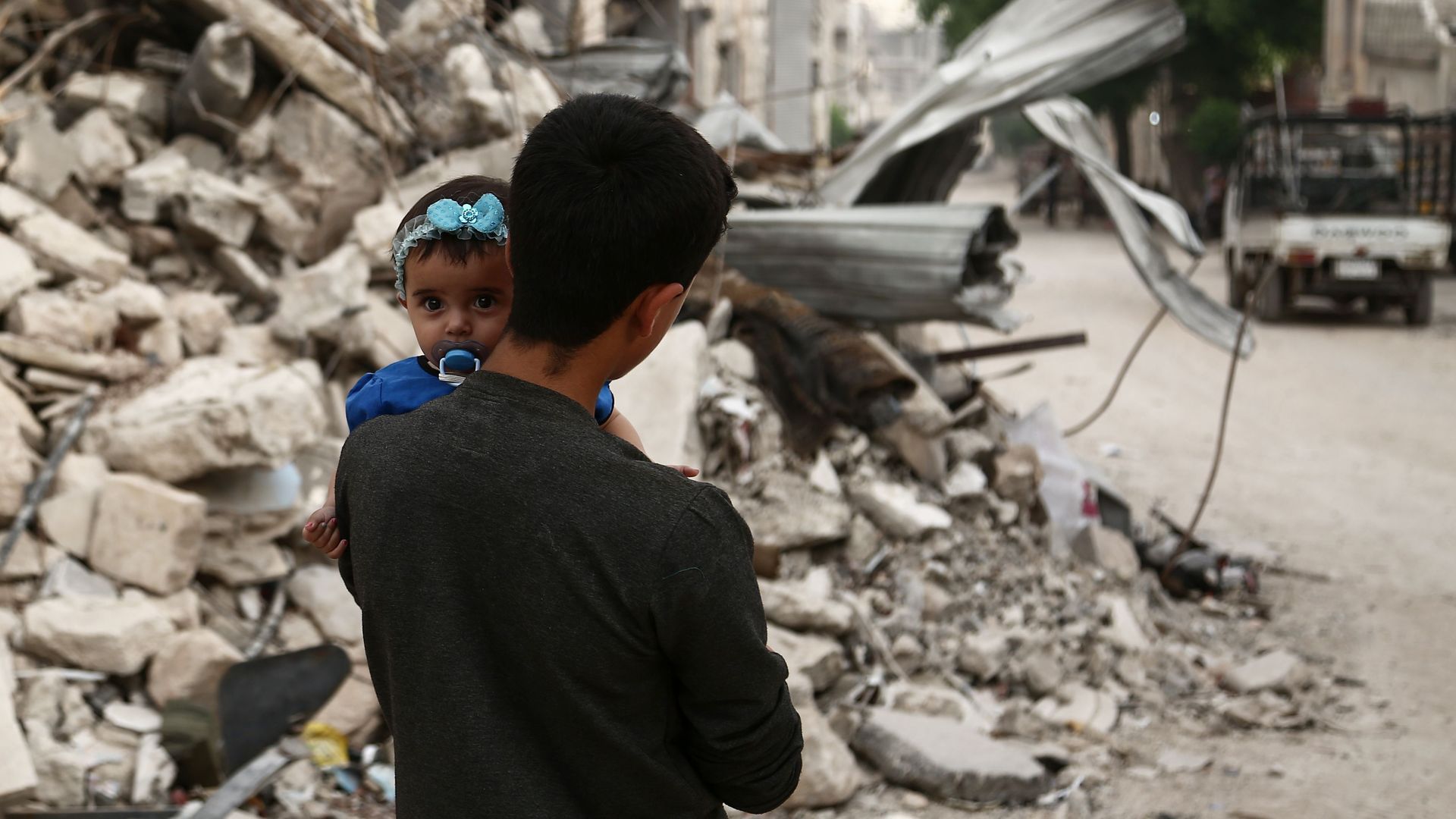 A Syrian youth walks by rubble holding a baby.