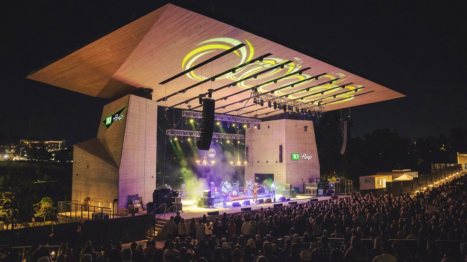 Nighttime outdoor concert on a large open-air stage with an angled wooden roof. Bright yellow circular light projections hang over a live band, as a large crowd watches from the front; TD logos on the towers.