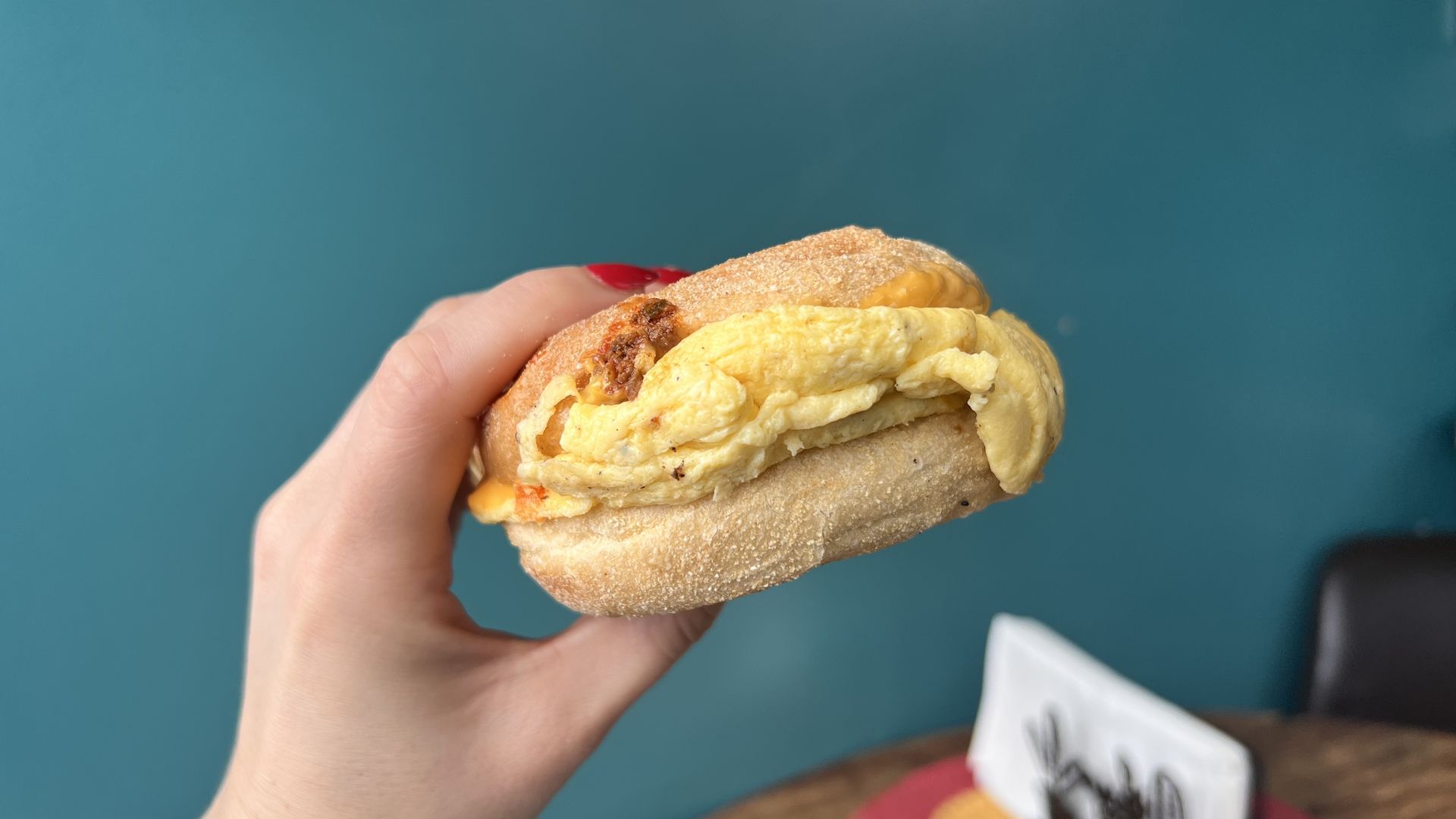 A picture of a hand witih red nail polish holding up a breakfast sandwich on an English muffin with yellow scrambled eggs showing. The background is a turquoise-ish wall.