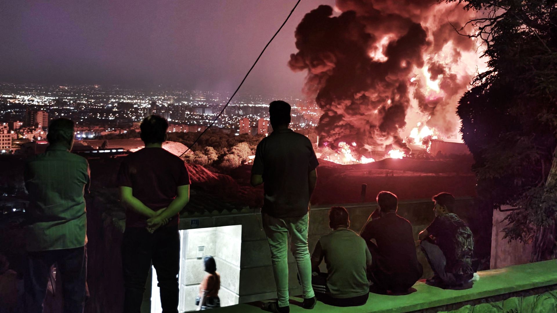 People observe fire and smoke from an Israeli attack on the Shahran oil depot on June 15, 2025 in Tehran, Iran.