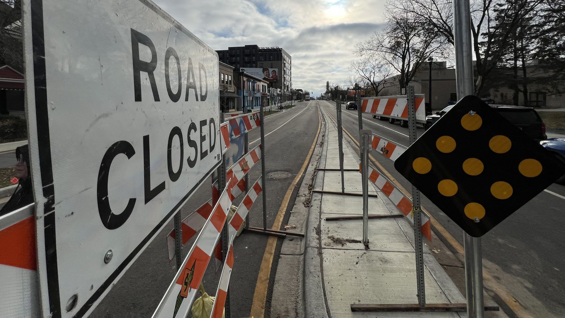 Urban construction site on a city street: a ROAD CLOSED sign and orange barricades flank rails in the center. Overcast sky with sun peeking through; bare trees and distant buildings.