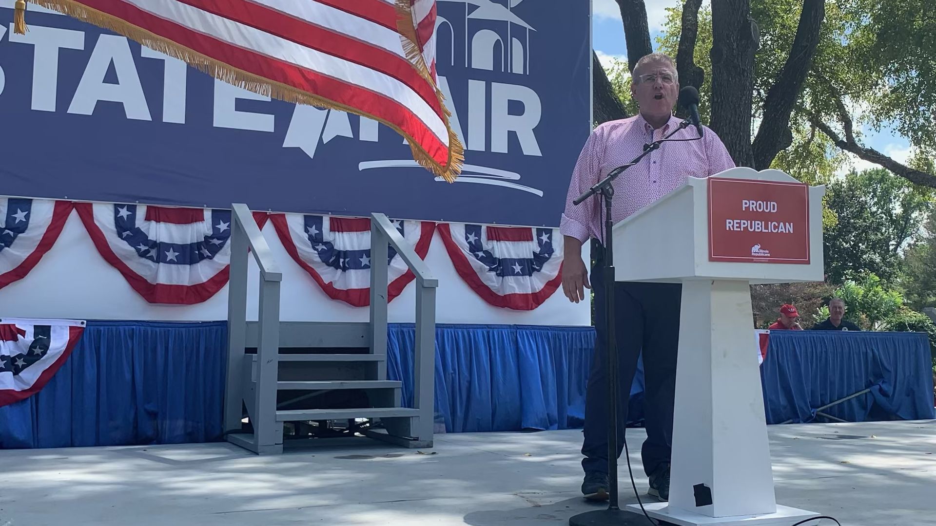 Outdoor political rally stage with a large American flag, a blue backdrop featuring a white building icon, red‑white‑blue bunting, and a man in a pink shirt speaking at a podium labeled PROUD REPUBLICAN.