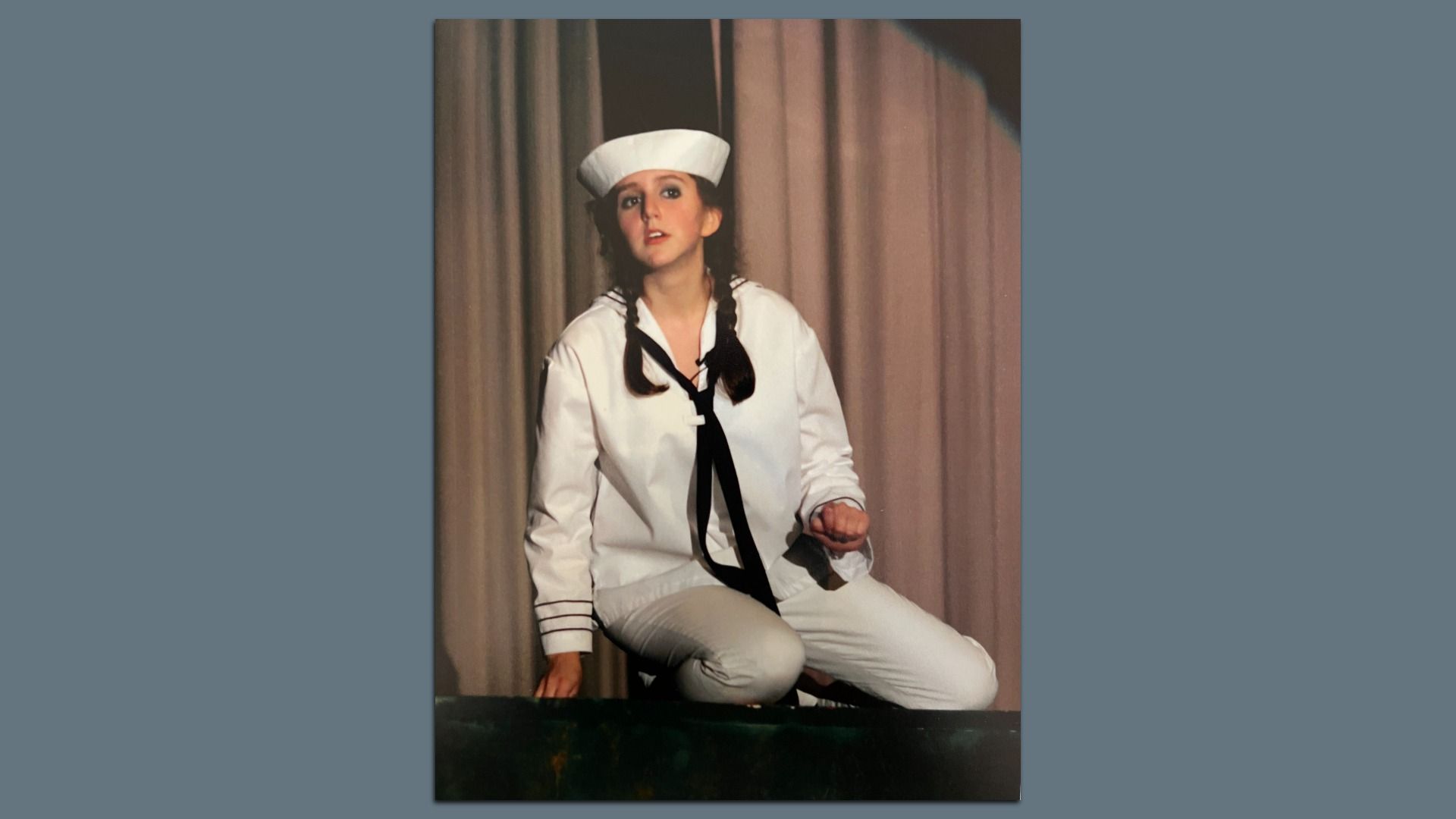 Young woman on stage in a white sailor costume, hat and black tie, with braided hair, seated before beige curtains, performing a scene.