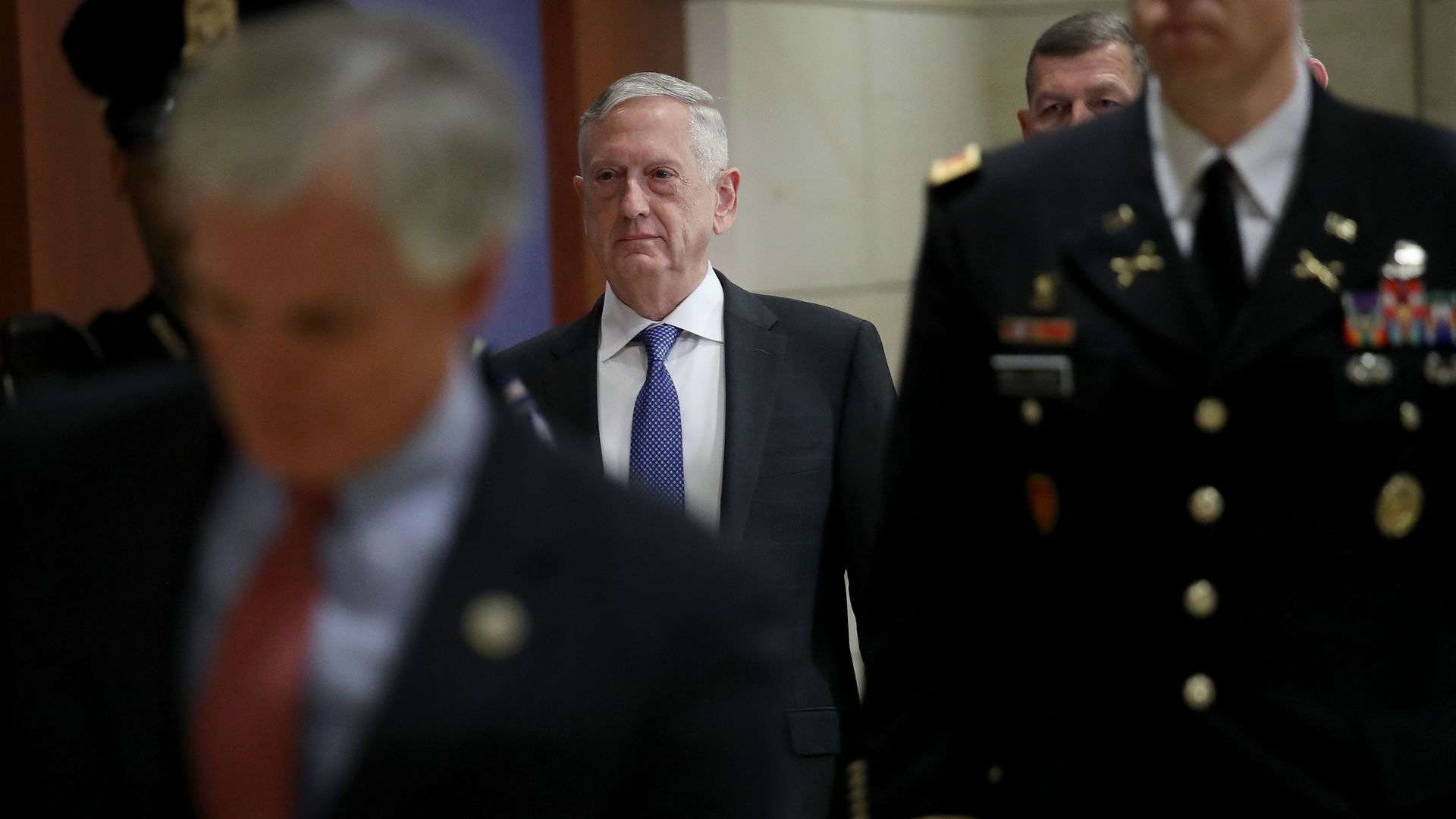 Jim Mattis walks down a hallway behind decorated and uniformed military personnel. 