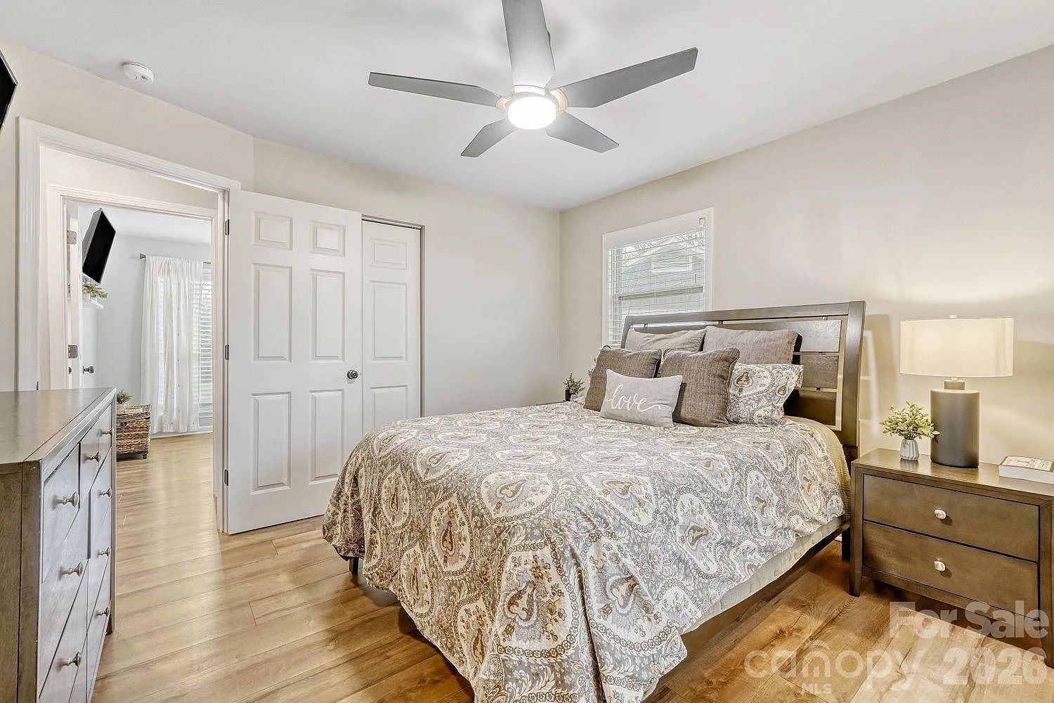 Bright bedroom with wooden floor, a bed with patterned gray and beige bedding, multiple pillows including one with "love" text, wooden dresser, nightstand with lamp and small plant, ceiling fan.