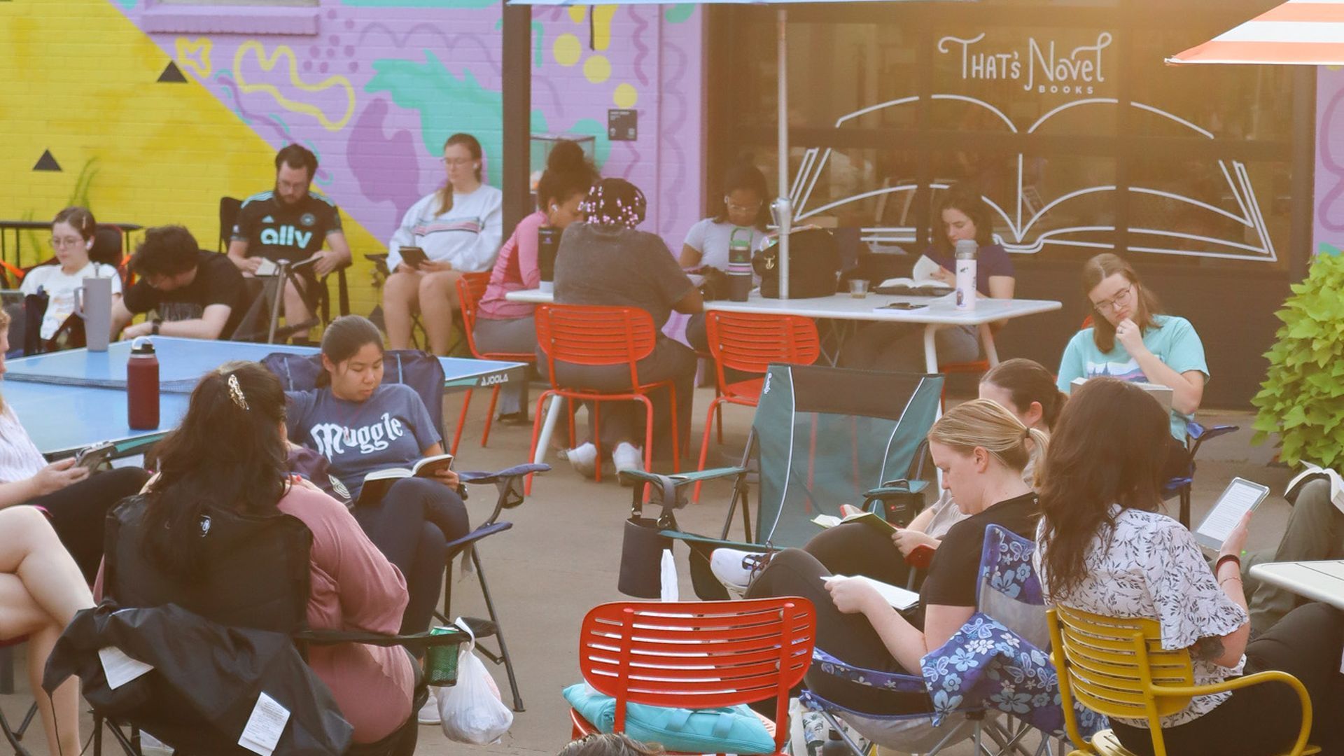 People sit around outdoor tables reading and chatting in front of a colorful mural; red and orange chairs surround a white table, and a sign reads "That's Novel Books".