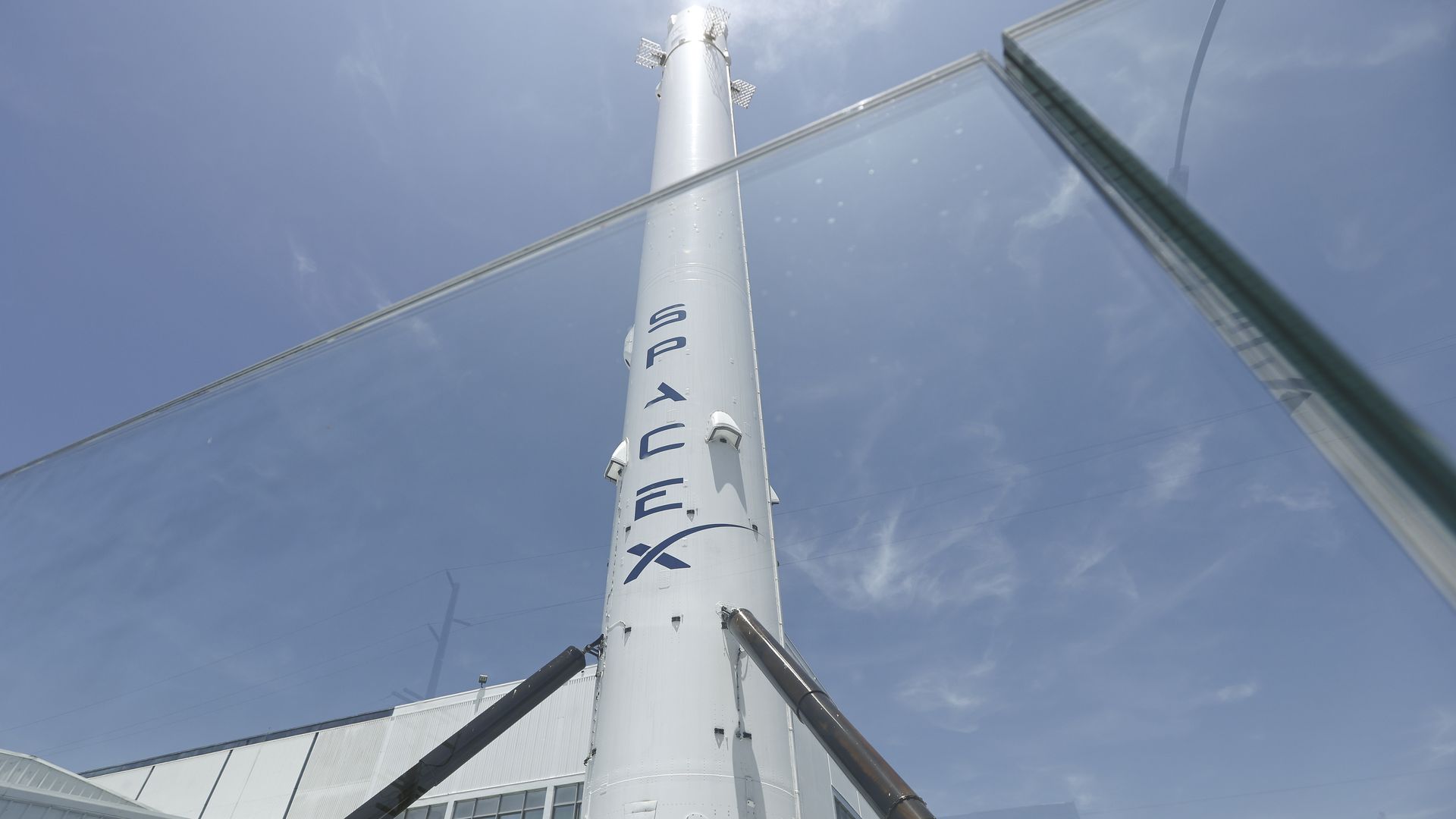 Tall white rocket-like column rises against a blue sky, seen through glass walls with diagonal metal supports; a modern building sits at the base and blue letters run vertically on the column.