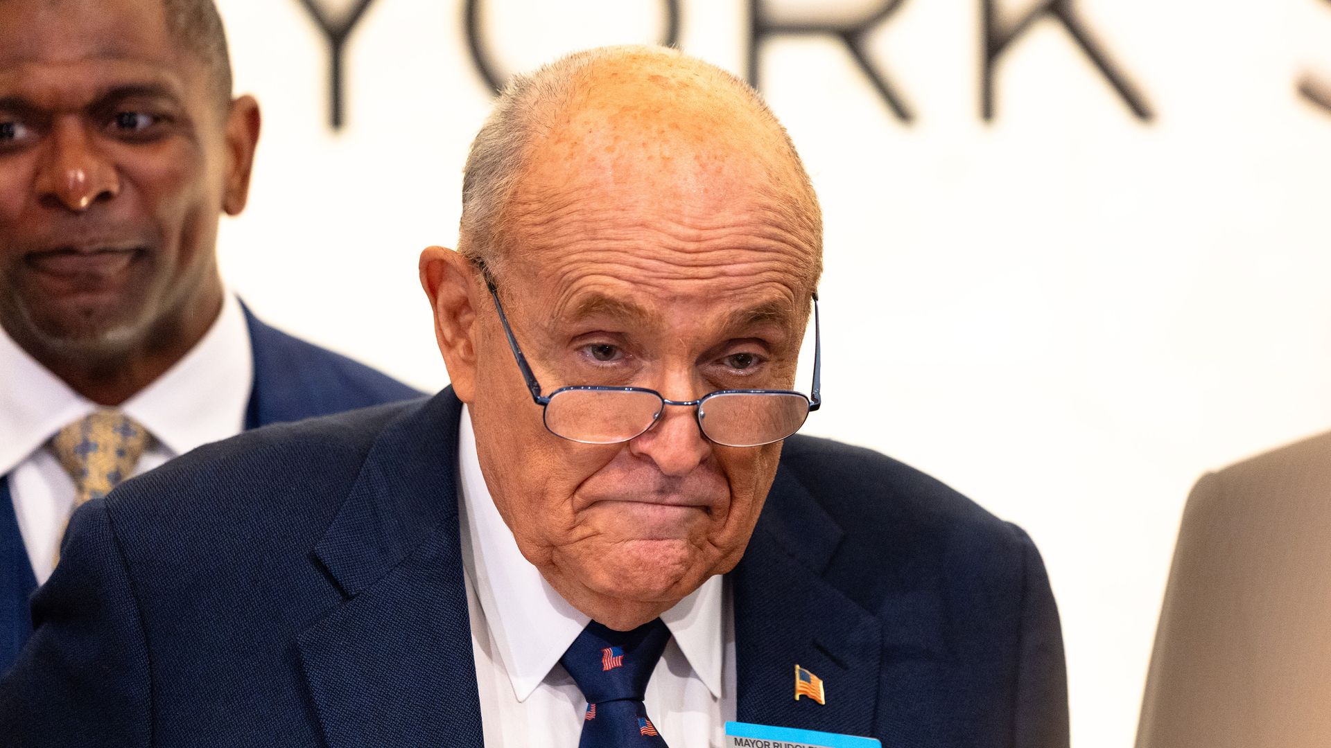 A bald, bespectacled former New York City Mayor Rudy Giuliani stares ahead while wearing a navy jacket with a US flag pin at the top of his left lapel, white shirt and navy tie with small US flags on it.
