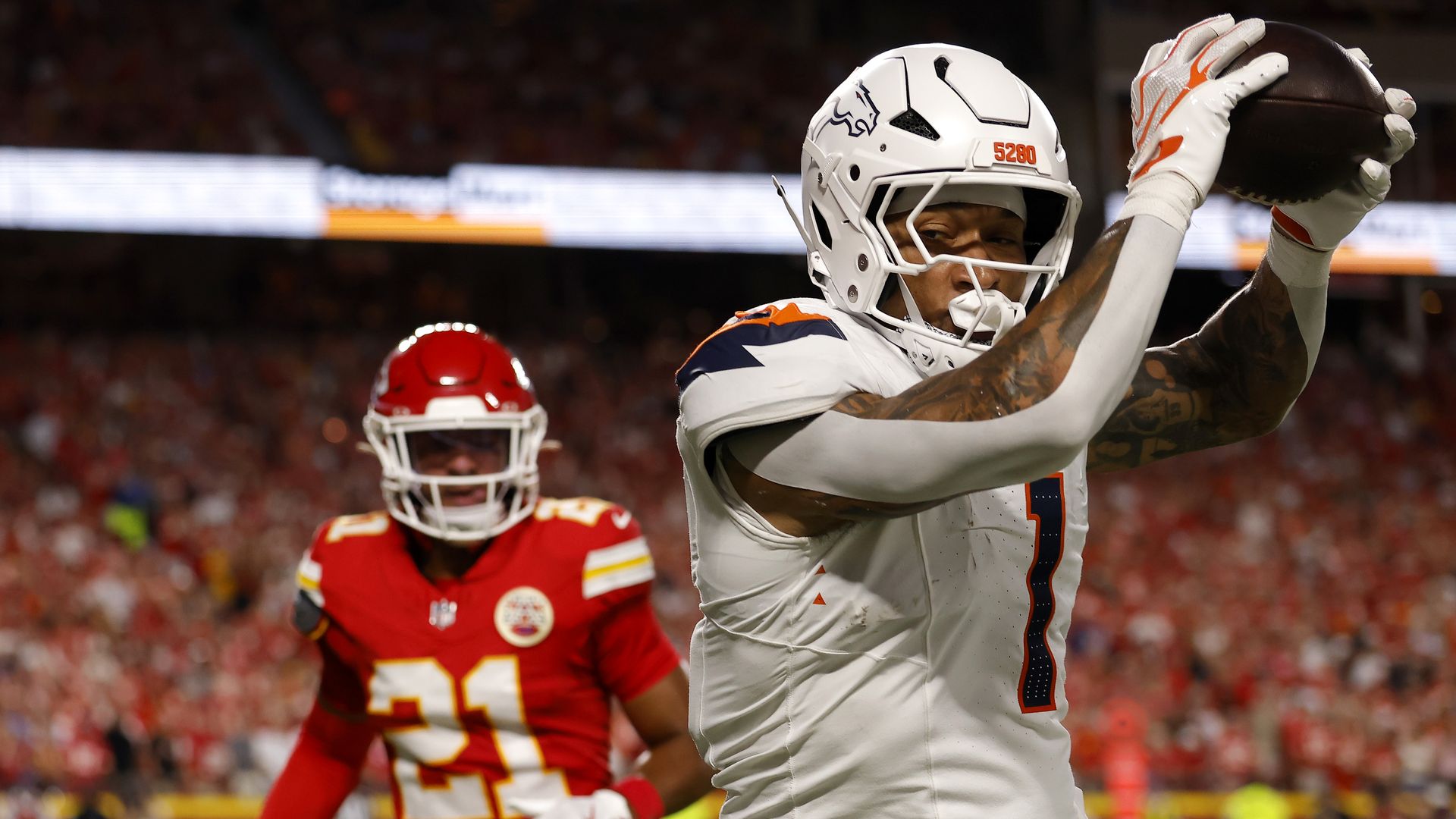Football player in a white uniform with orange and blue trim, number 7, raises a football overhead in celebration as a red-uniform defender closes in; stadium crowd blurred in background.